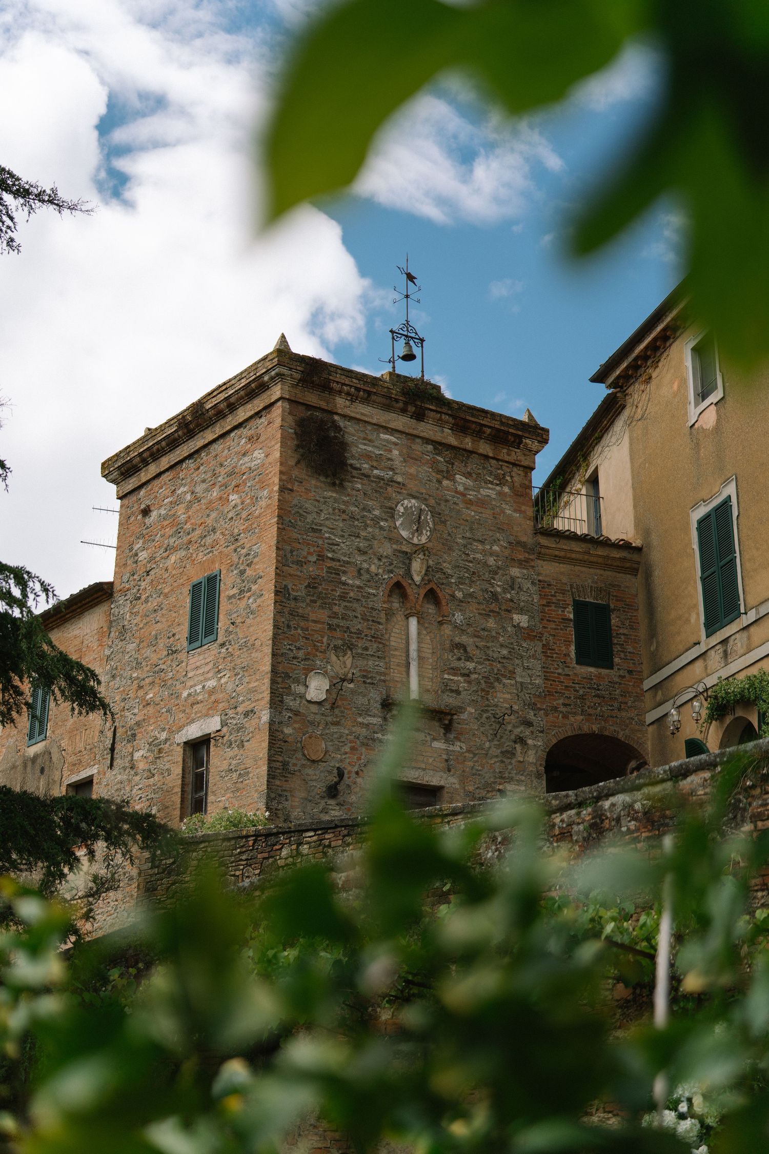 Historic medieval stone tower with clock face framed by green foliage against a blue cloudy sky.