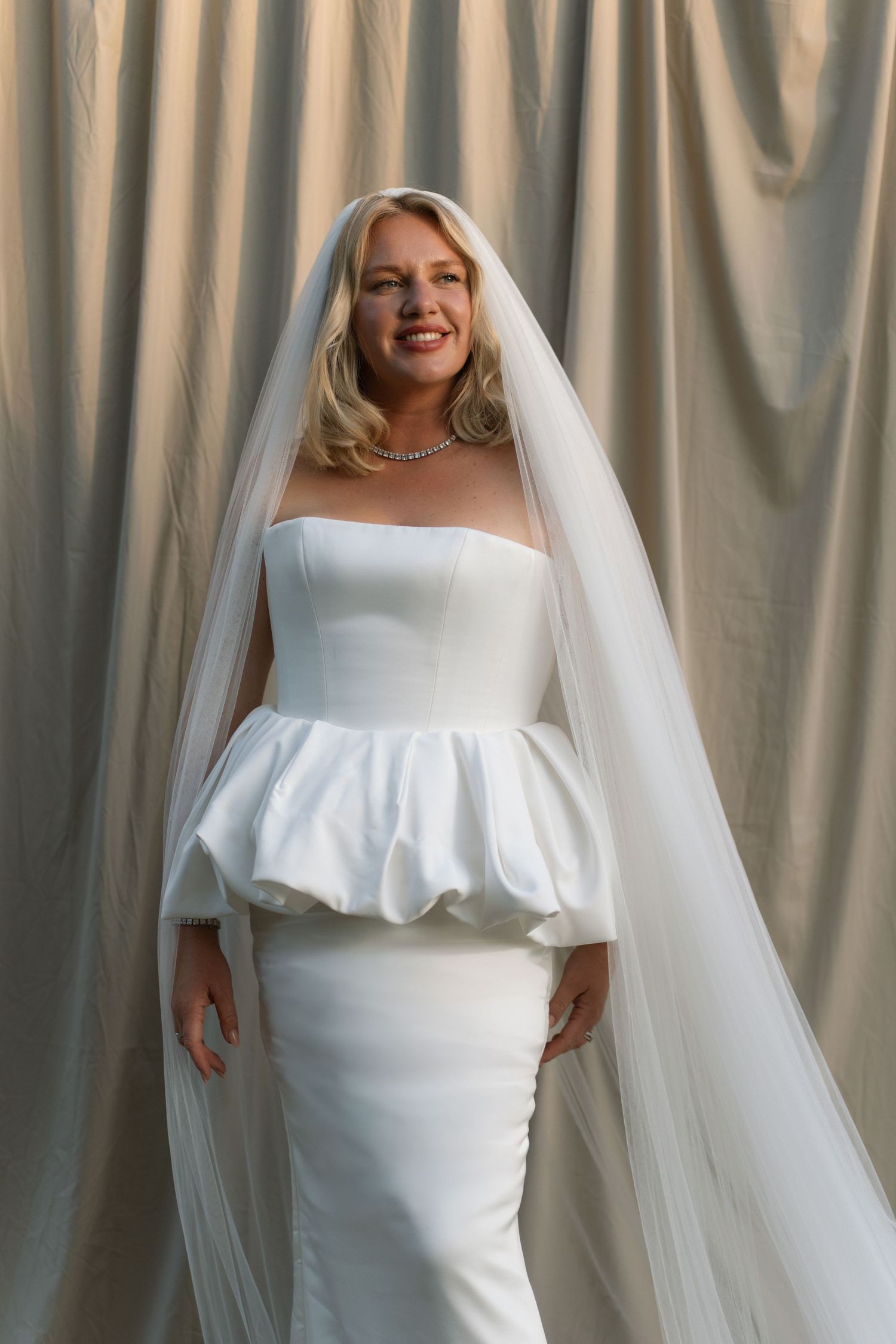 Bride in strapless white peplum wedding gown with long veil and pearl necklace, posing against draped fabric backdrop.