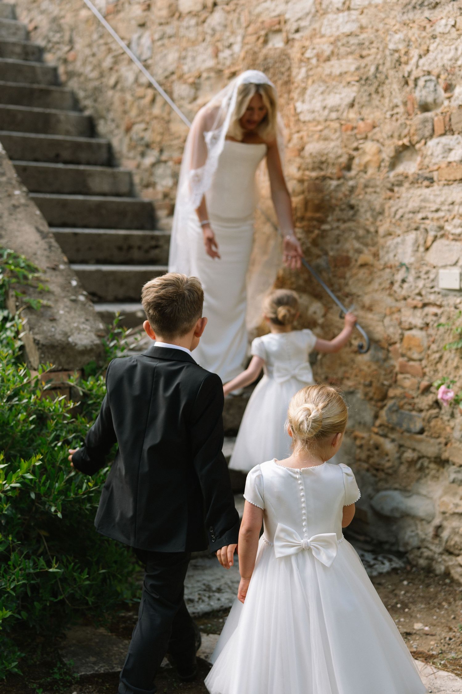 Bride in white gown and veil guides children in formal attire up stone steps at a wedding venue.