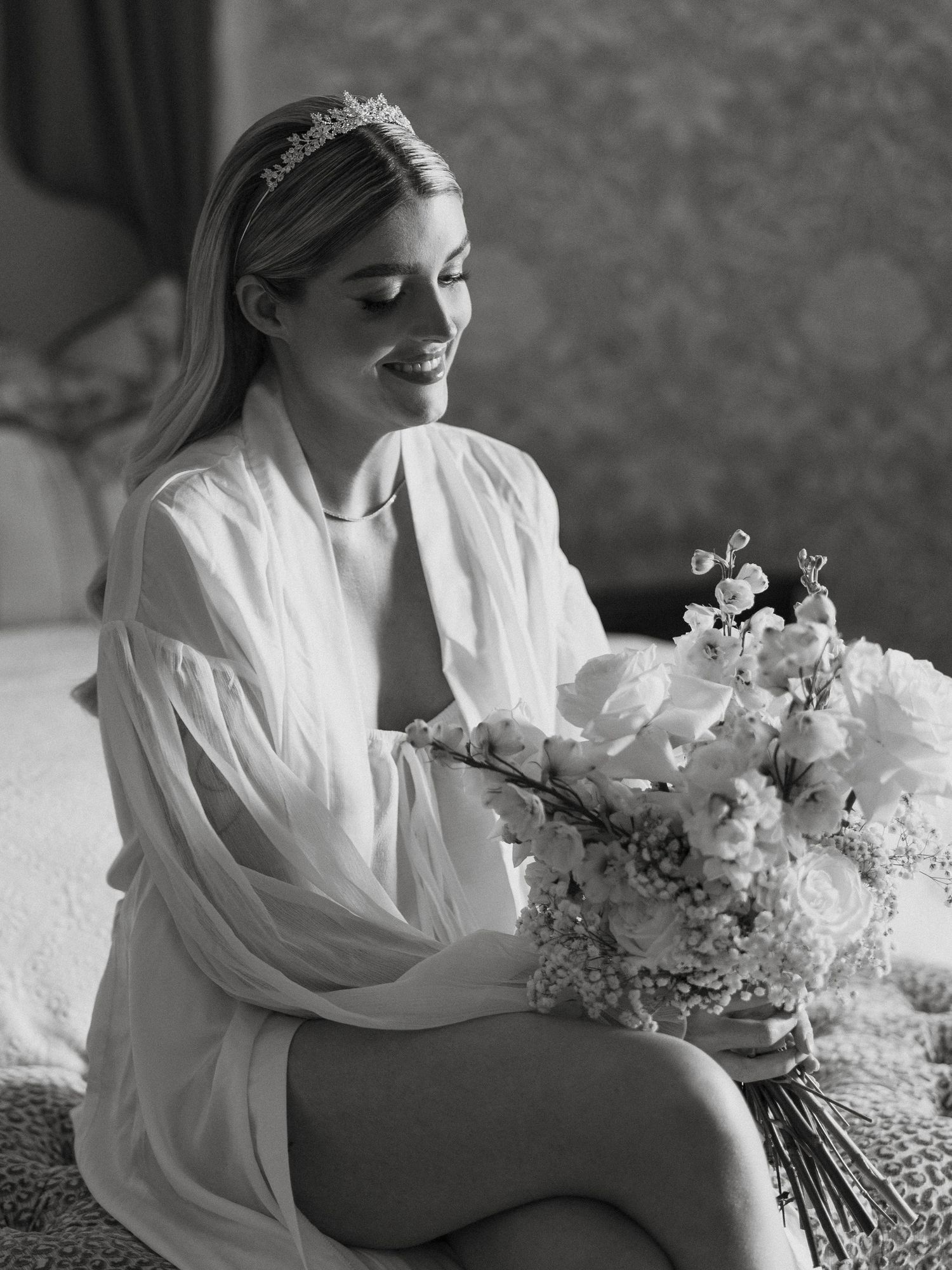 Black and white photo of a smiling bride in a sheer robe wearing a tiara, holding a bouquet of flowers.