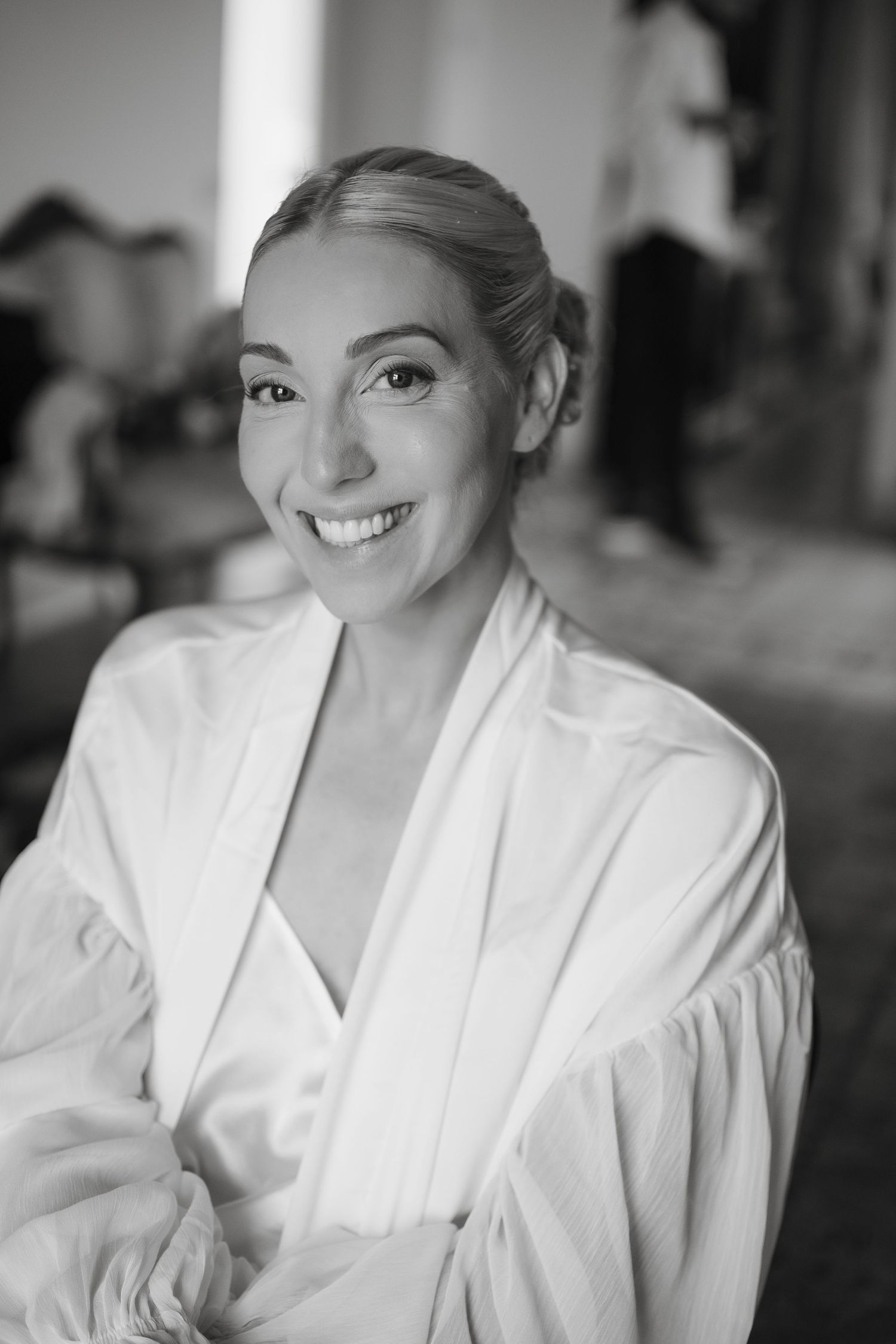 Black and white portrait of a smiling woman in a white robe, elegantly posed in a softly lit room.