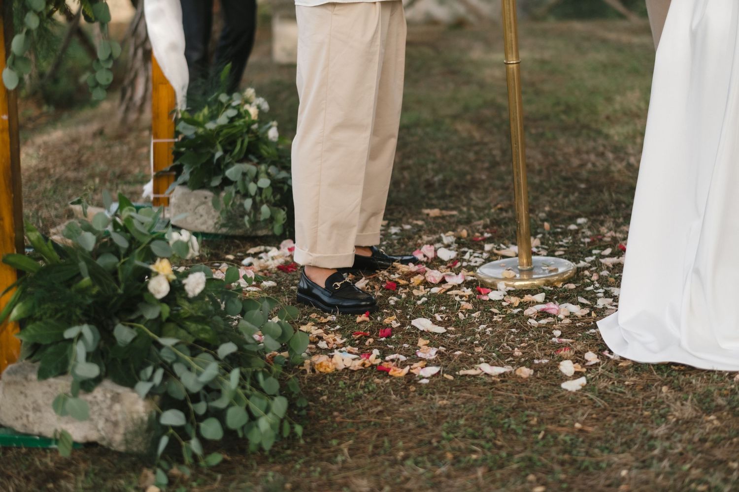 Outdoor wedding ceremony close-up showing feet of couple standing on petal-strewn ground with floral greenery arch.