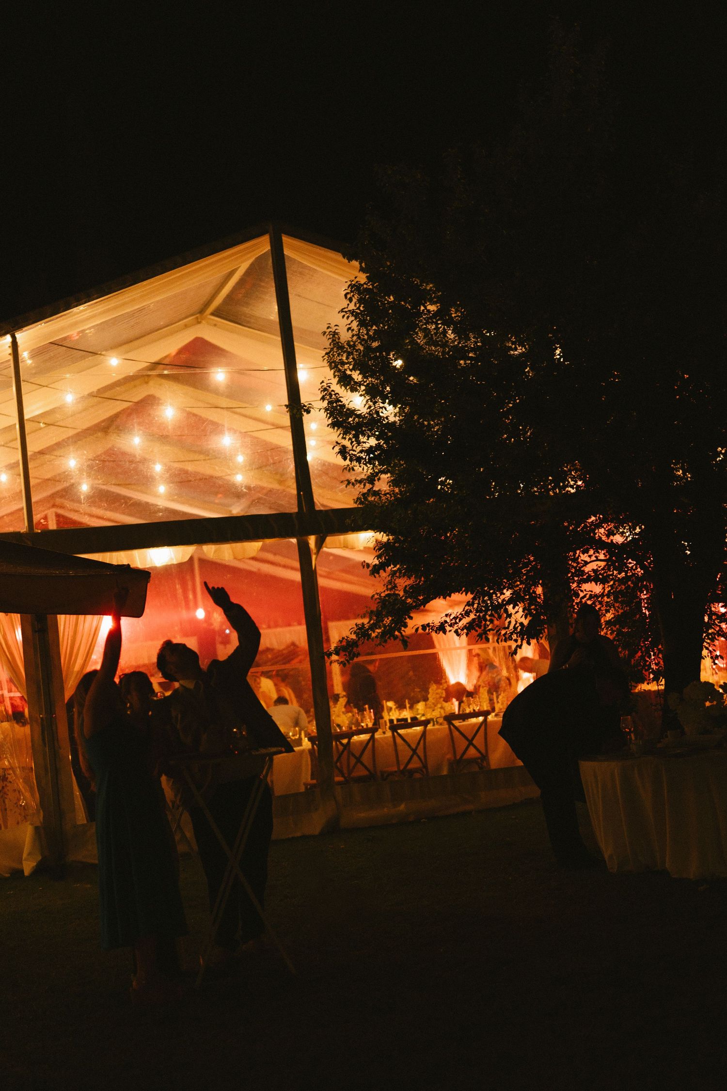 Illuminated wedding reception tent glowing warmly at night with silhouettes of guests outside.