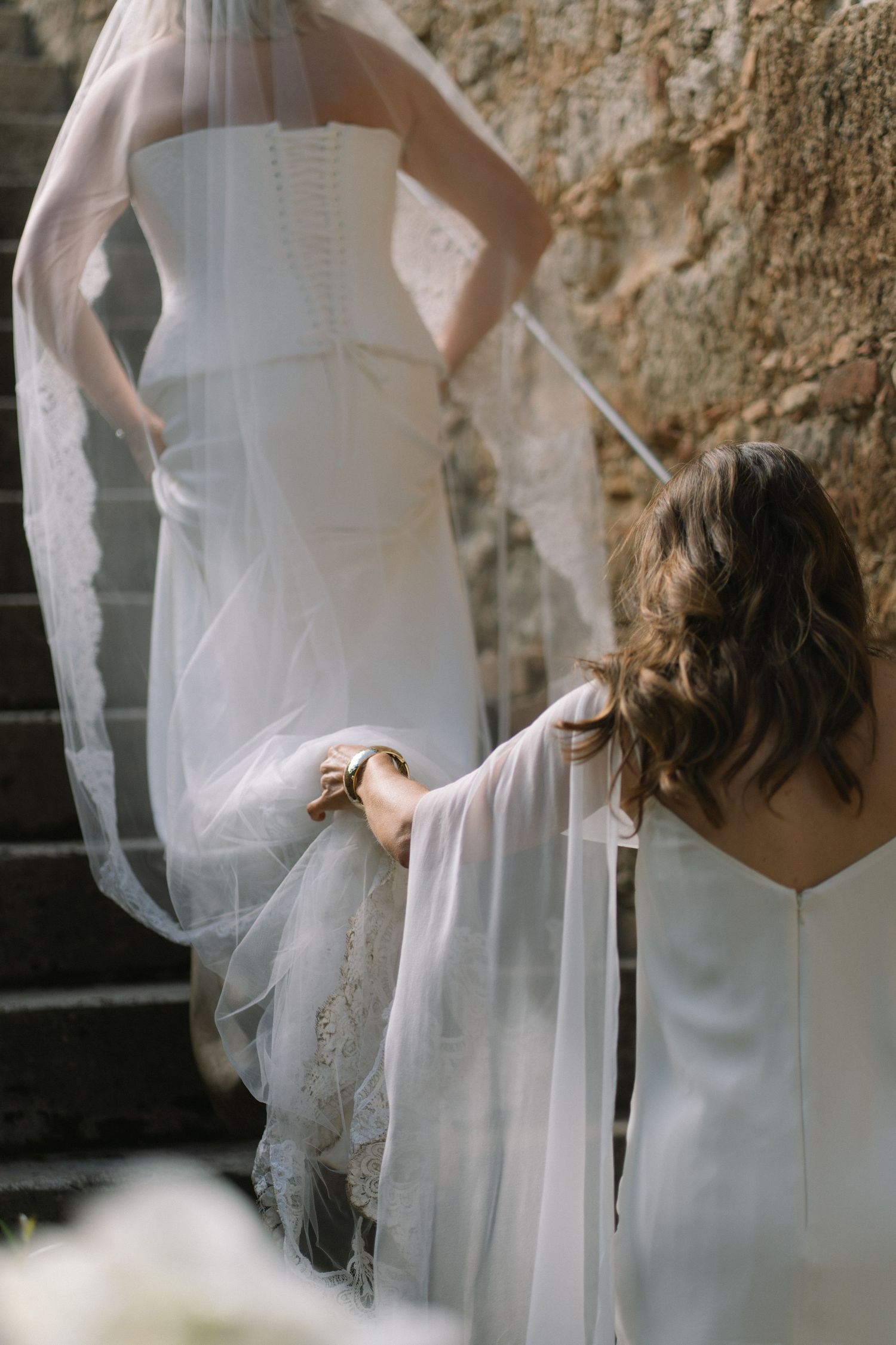 Bride in white lace gown ascending stone steps with bridesmaid holding her veil and train.