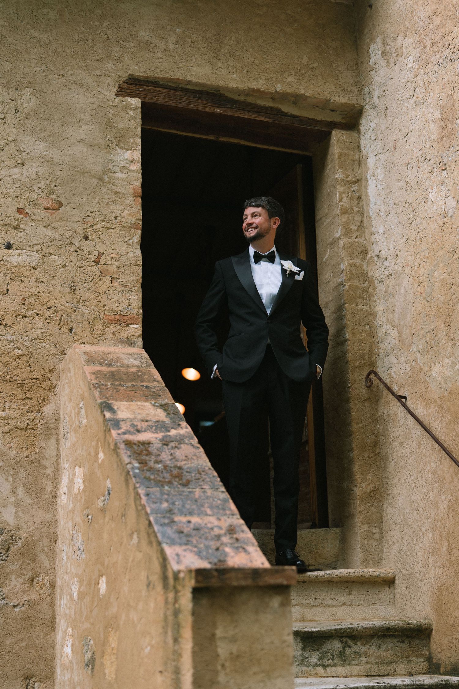 Handsome groom in black tuxedo with boutonniere standing in doorway of rustic stone building staircase.