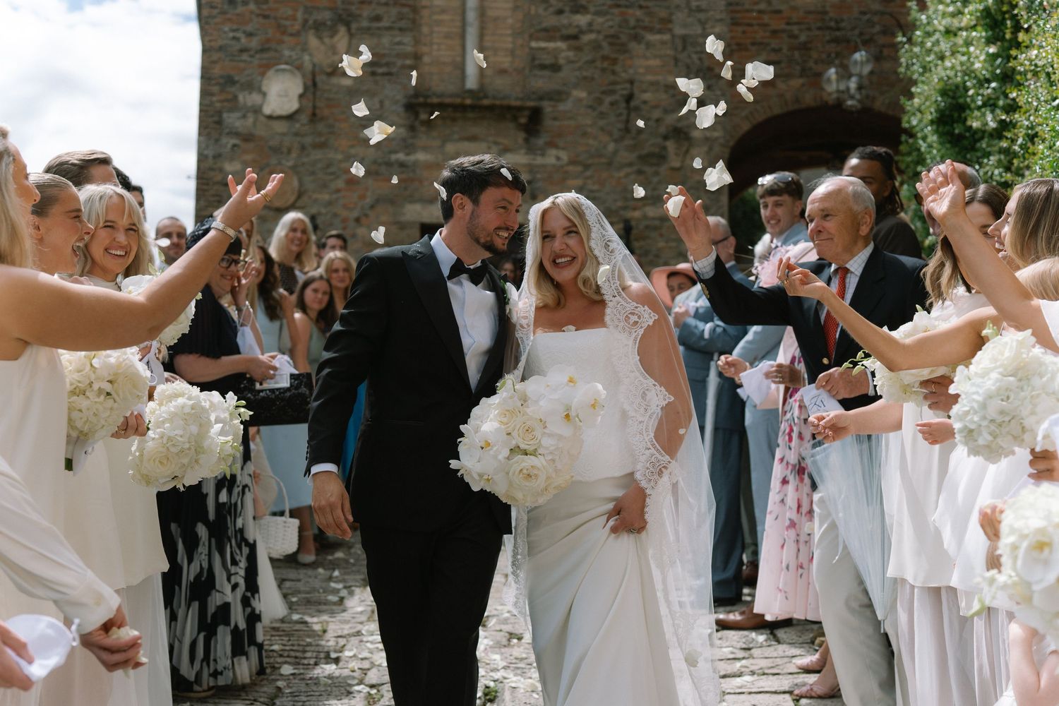 Newlyweds walk through a confetti shower surrounded by cheering guests after their wedding ceremony.