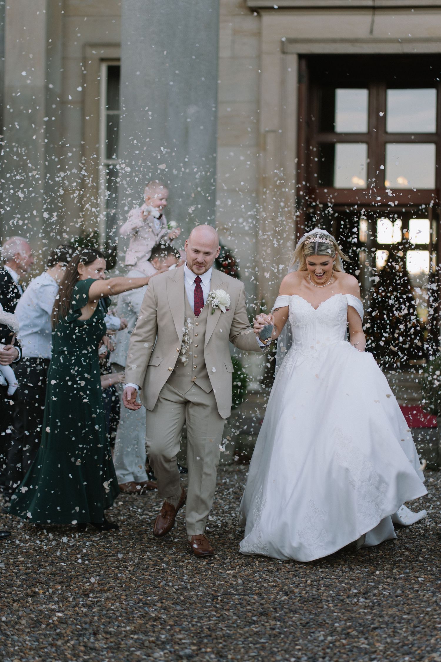 Bride and groom walk through confetti shower outside elegant venue, groom in tan suit, bride in white off-shoulder gown.
