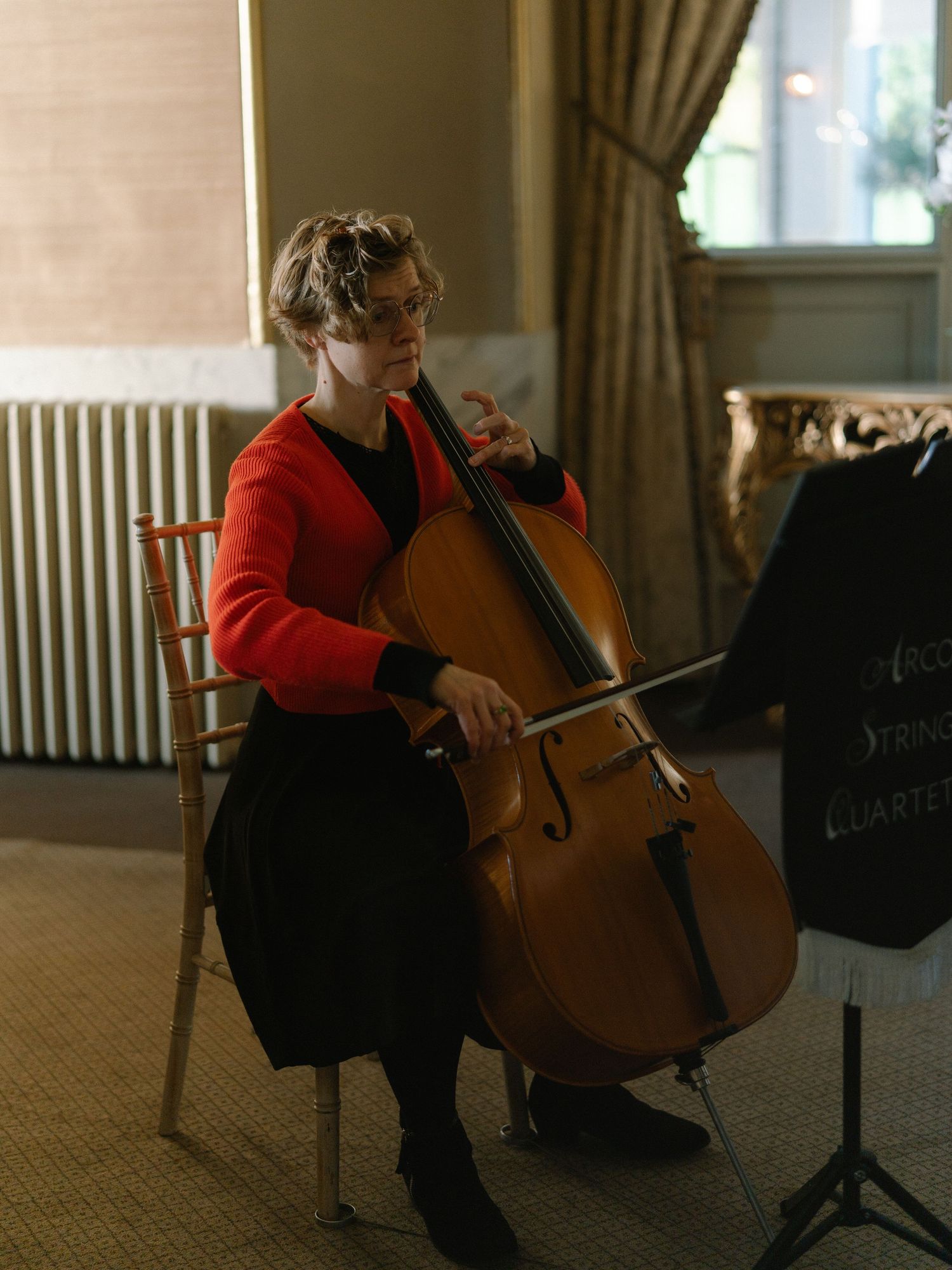 A cellist in a red cardigan plays cello while reading sheet music on a stand in an elegant room.