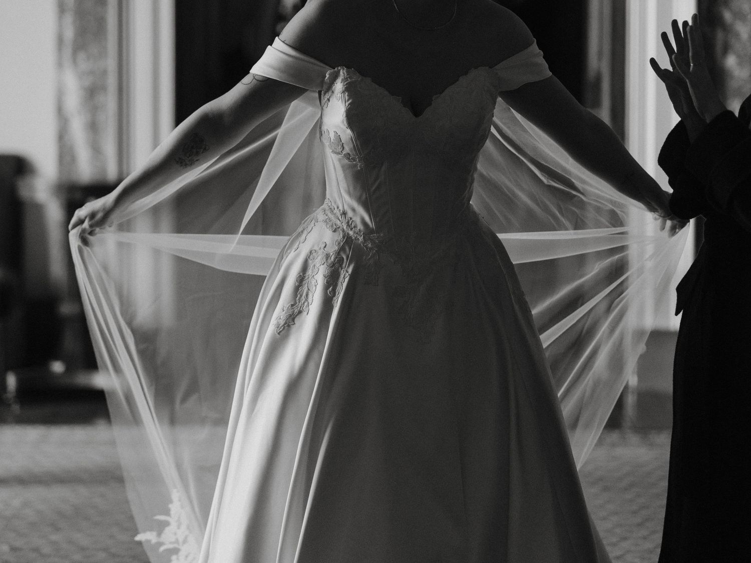 Black and white photo of a bride in an off-shoulder wedding gown holding her veil wide open.