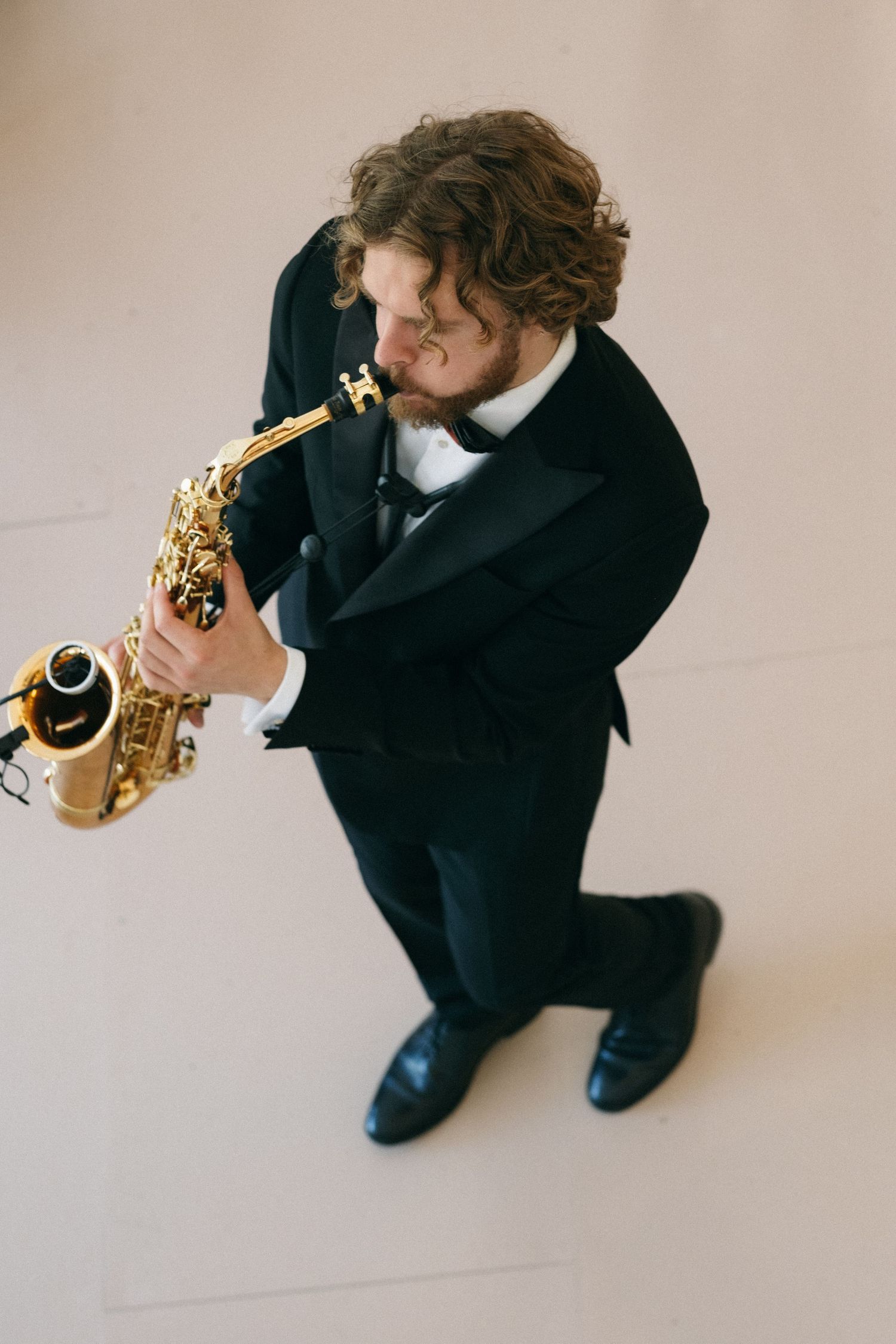 Aerial view of a male saxophonist in a black tuxedo playing a gold saxophone on a light floor.