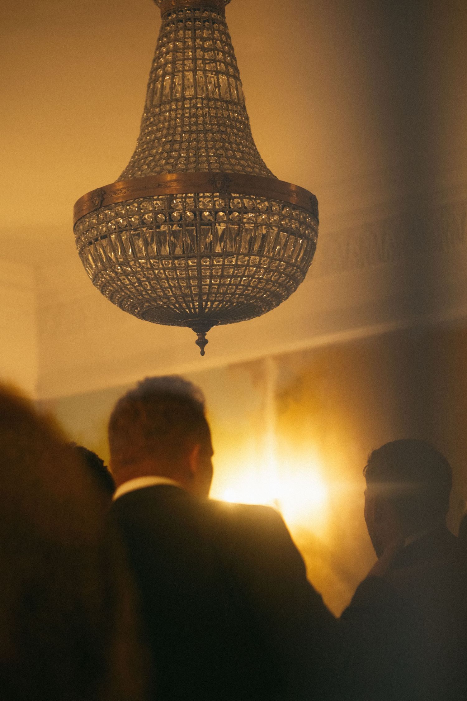 Elegant crystal chandelier hanging above silhouetted guests at a formal event with warm golden light.