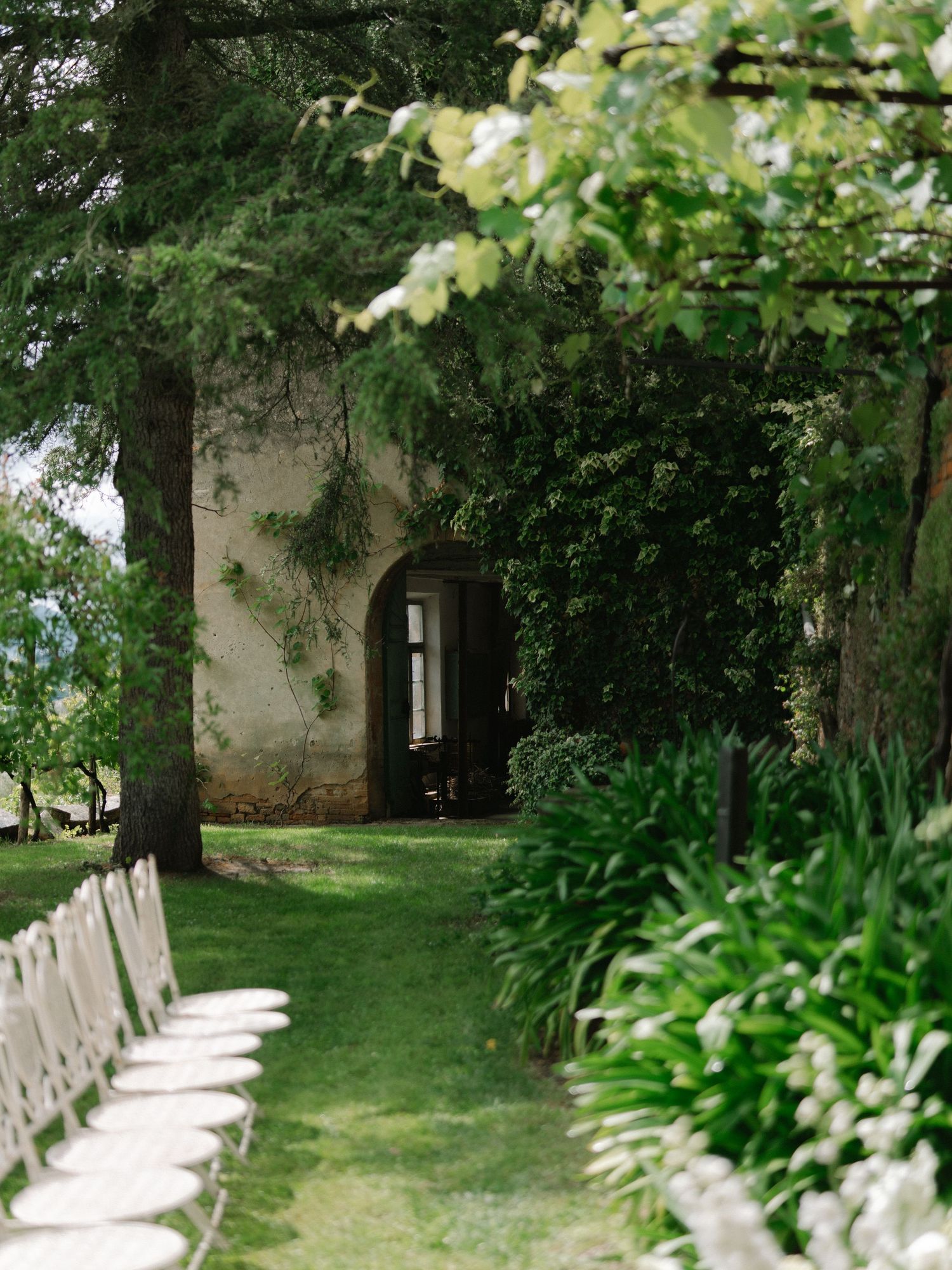 White chairs lining grass aisle leading to rustic stone chapel surrounded by tall trees.