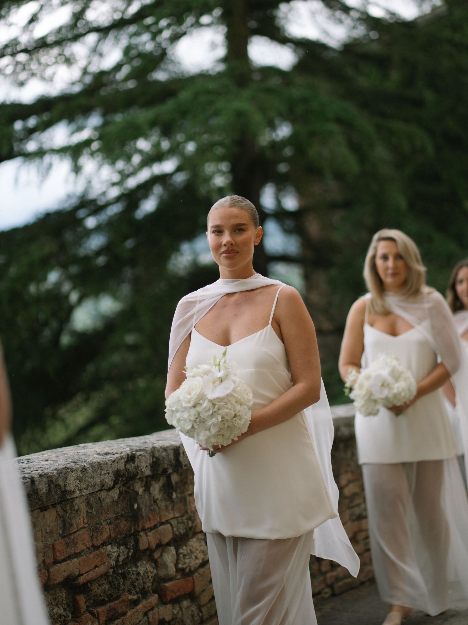 Elegant bridesmaid with shaved head in white draped gown carrying white hydrangea bouquet at outdoor wedding.