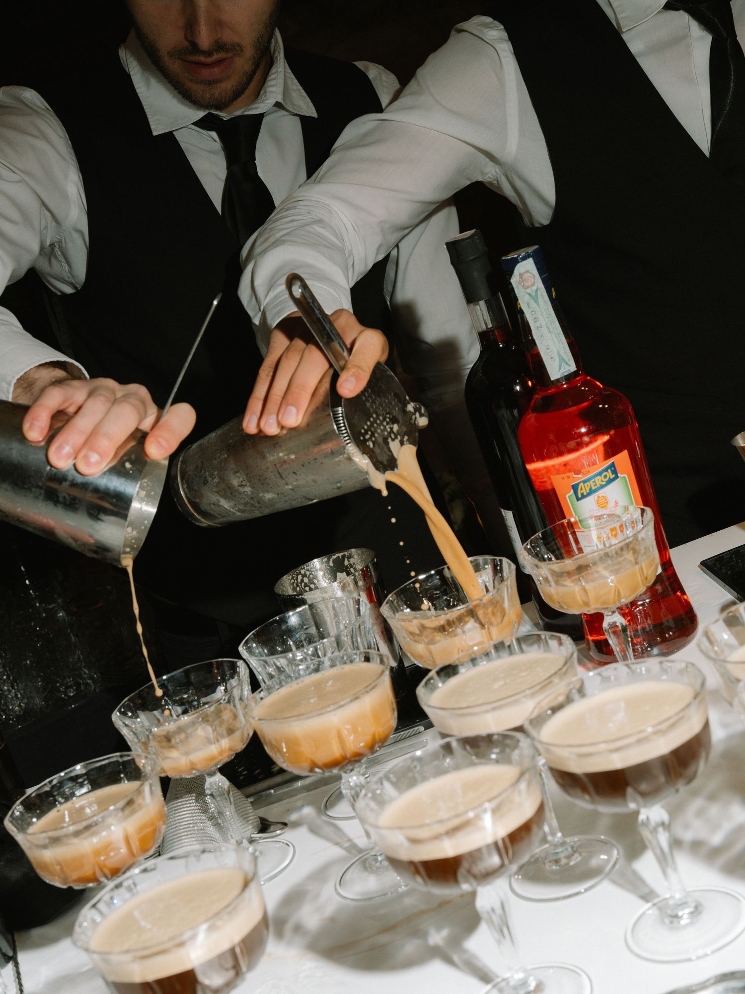 Bartender pouring espresso martinis into coupe glasses with Aperol bottle at wedding bar.