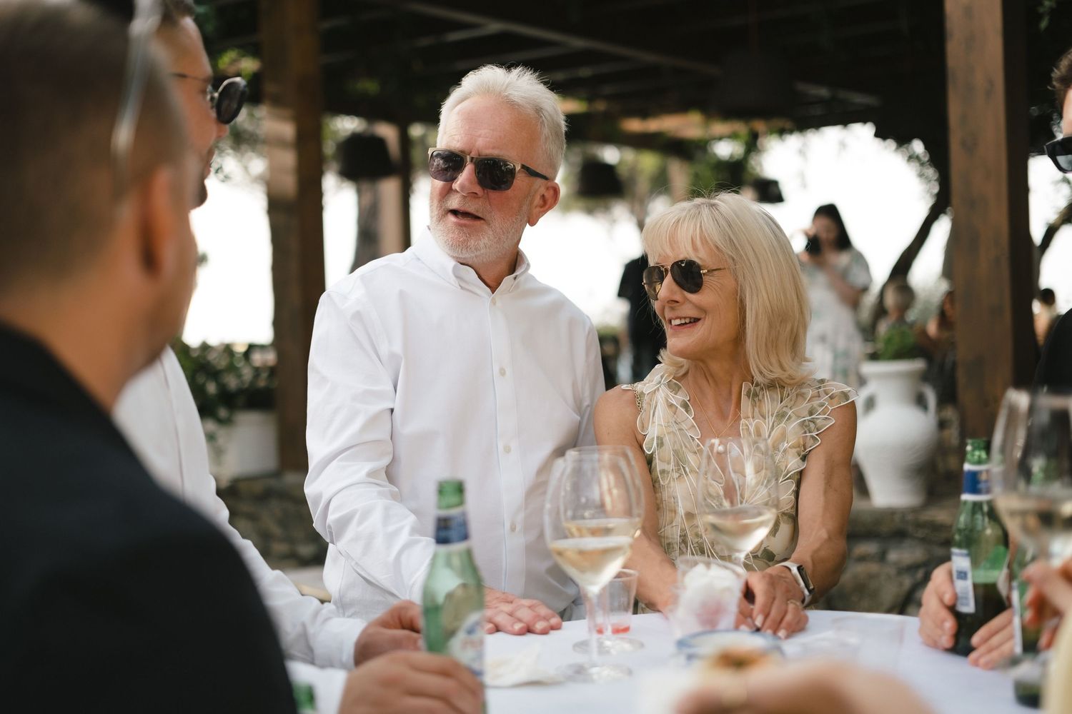 Elegant senior couple in sunglasses smiling at an outdoor wedding reception dinner table with wine glasses.