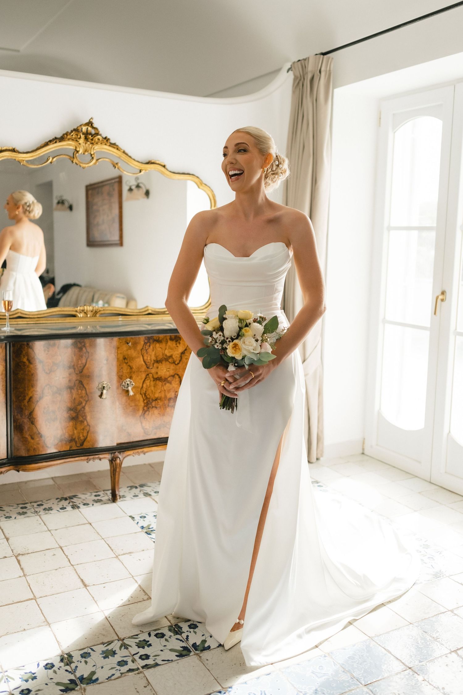 Radiant blonde bride in strapless white gown with slit holding bouquet indoors.