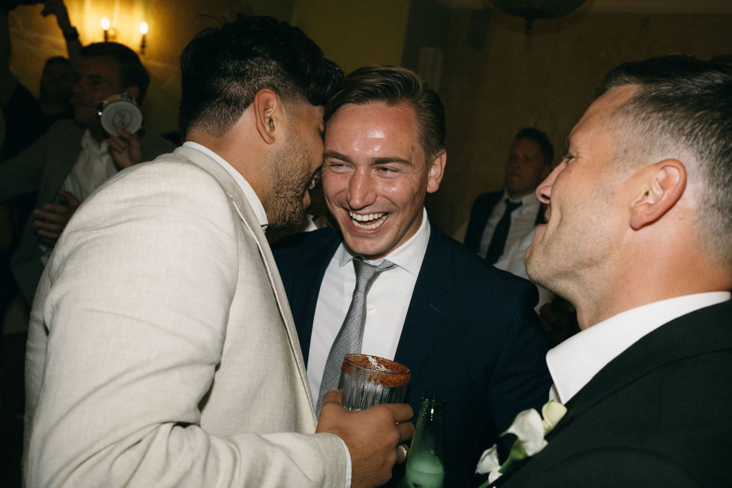 Three men in formal attire laughing and celebrating together at a wedding reception.