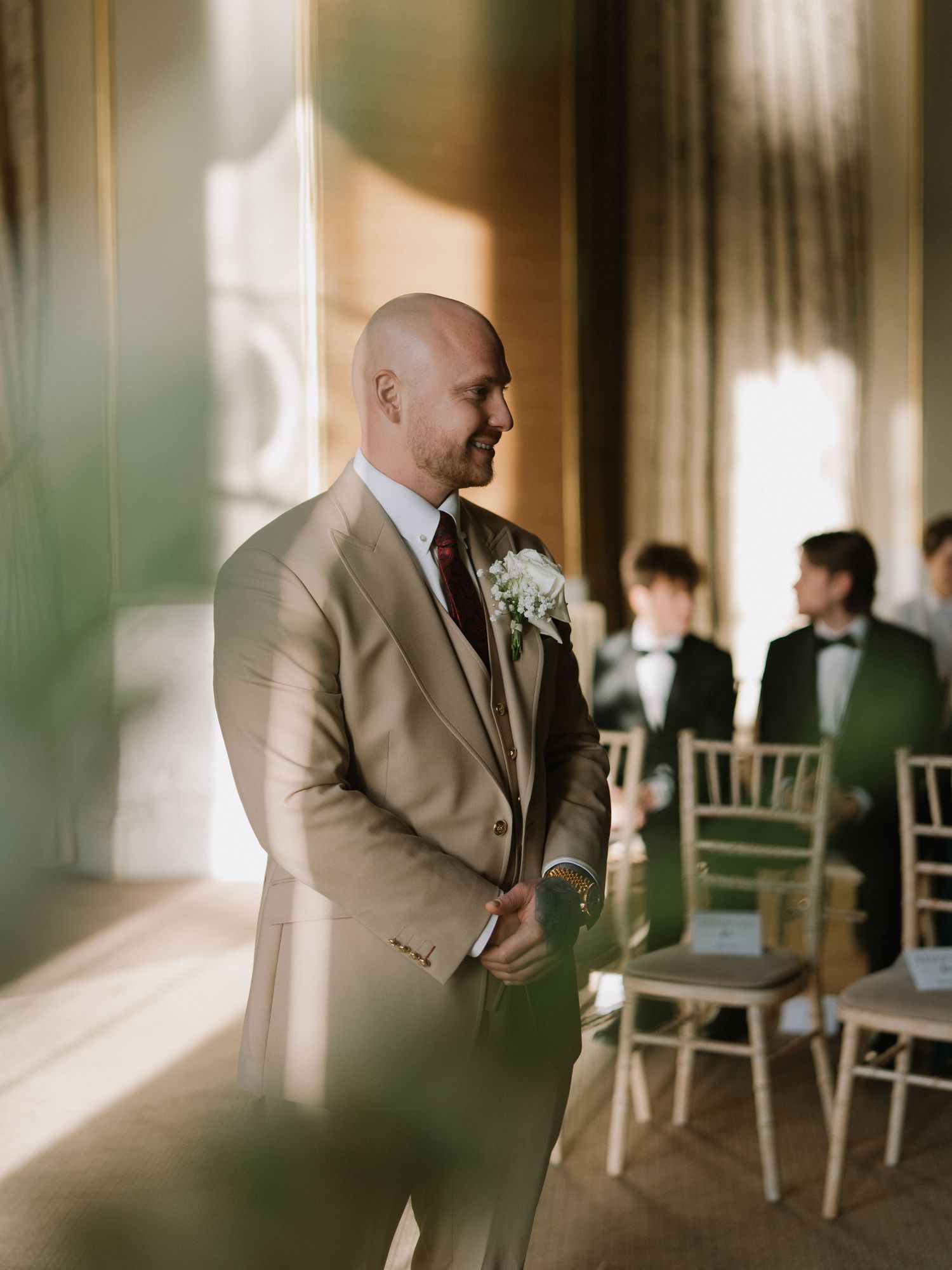 Bald groom in tan suit with white boutonniere stands at wedding ceremony altar in elegant venue.