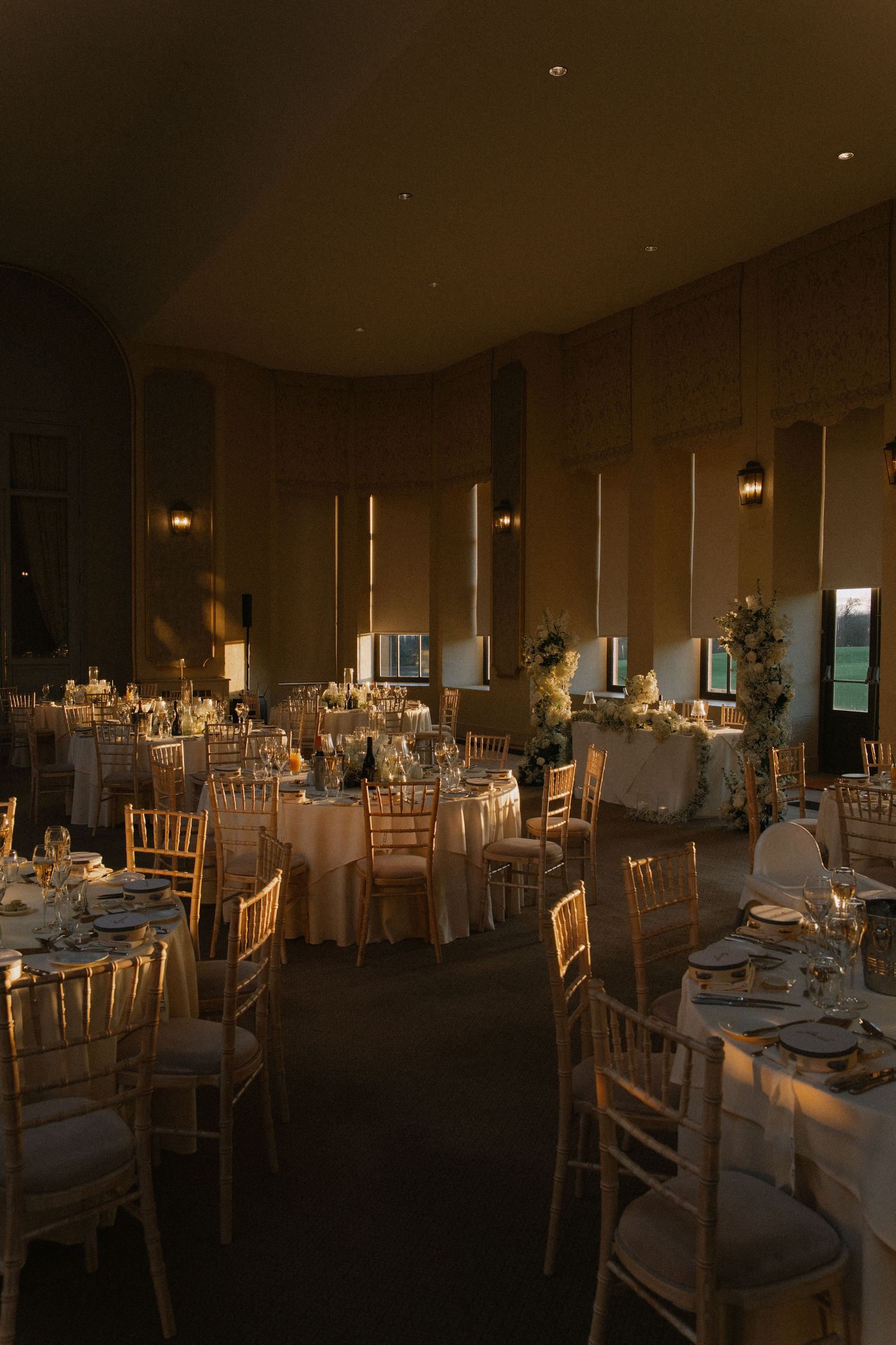 Elegant wedding reception hall with gold chiavari chairs, round tables, white linens, and warm ambient lighting.