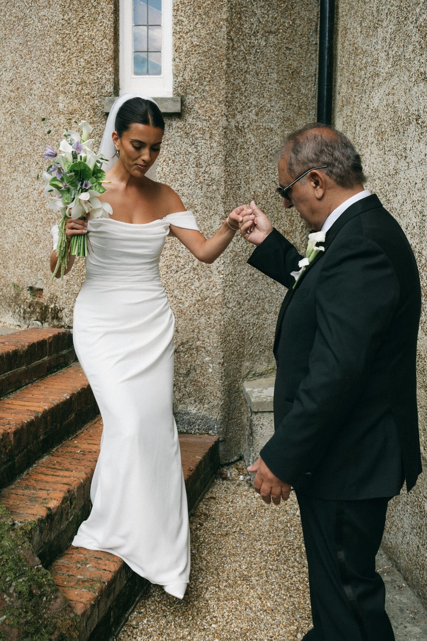 Bride in white off-shoulder gown holding bouquet, guided down steps by older man in black suit.