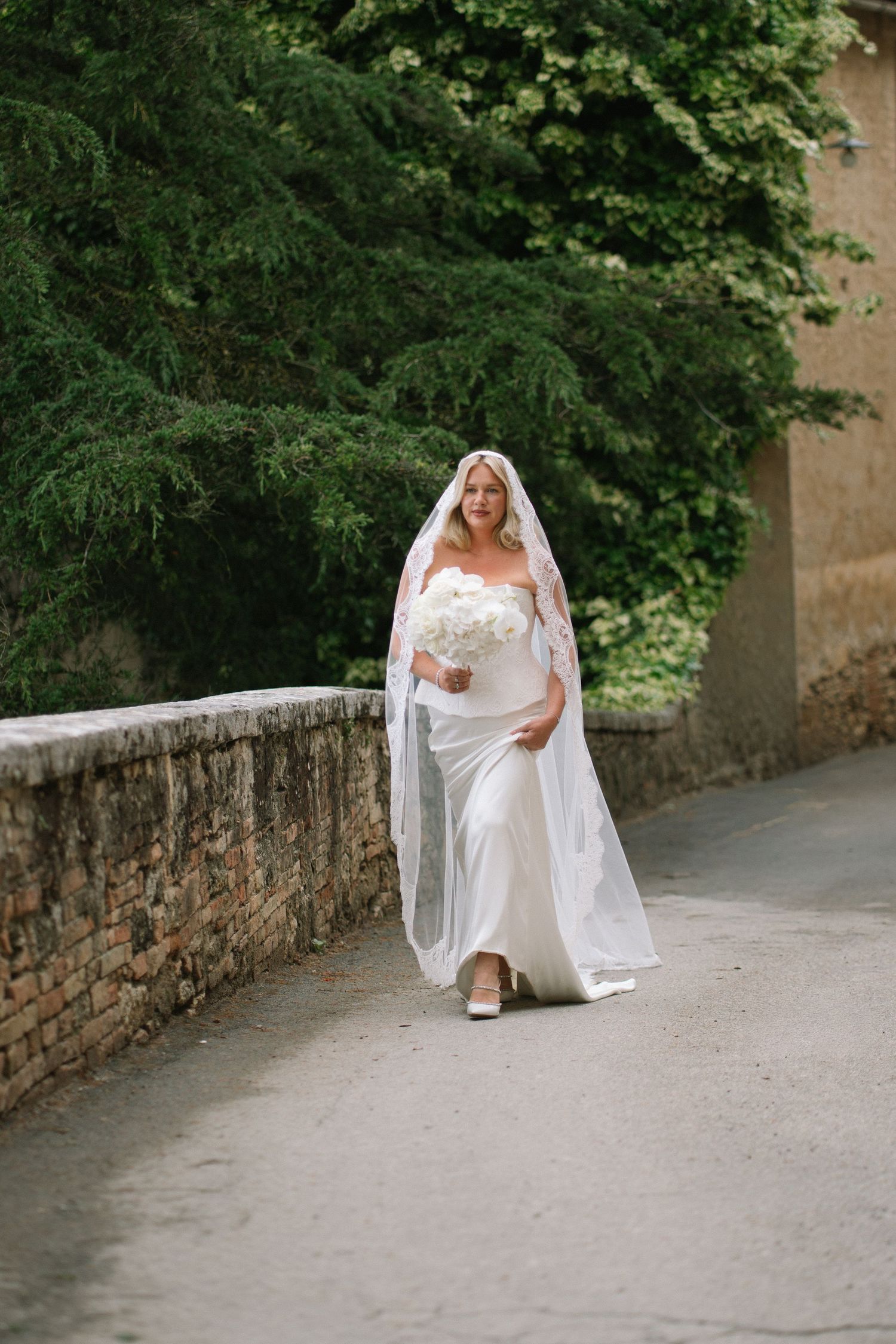 Radiant bride in fitted white gown with long lace-edged veil carrying white bouquet walking along a stone path.