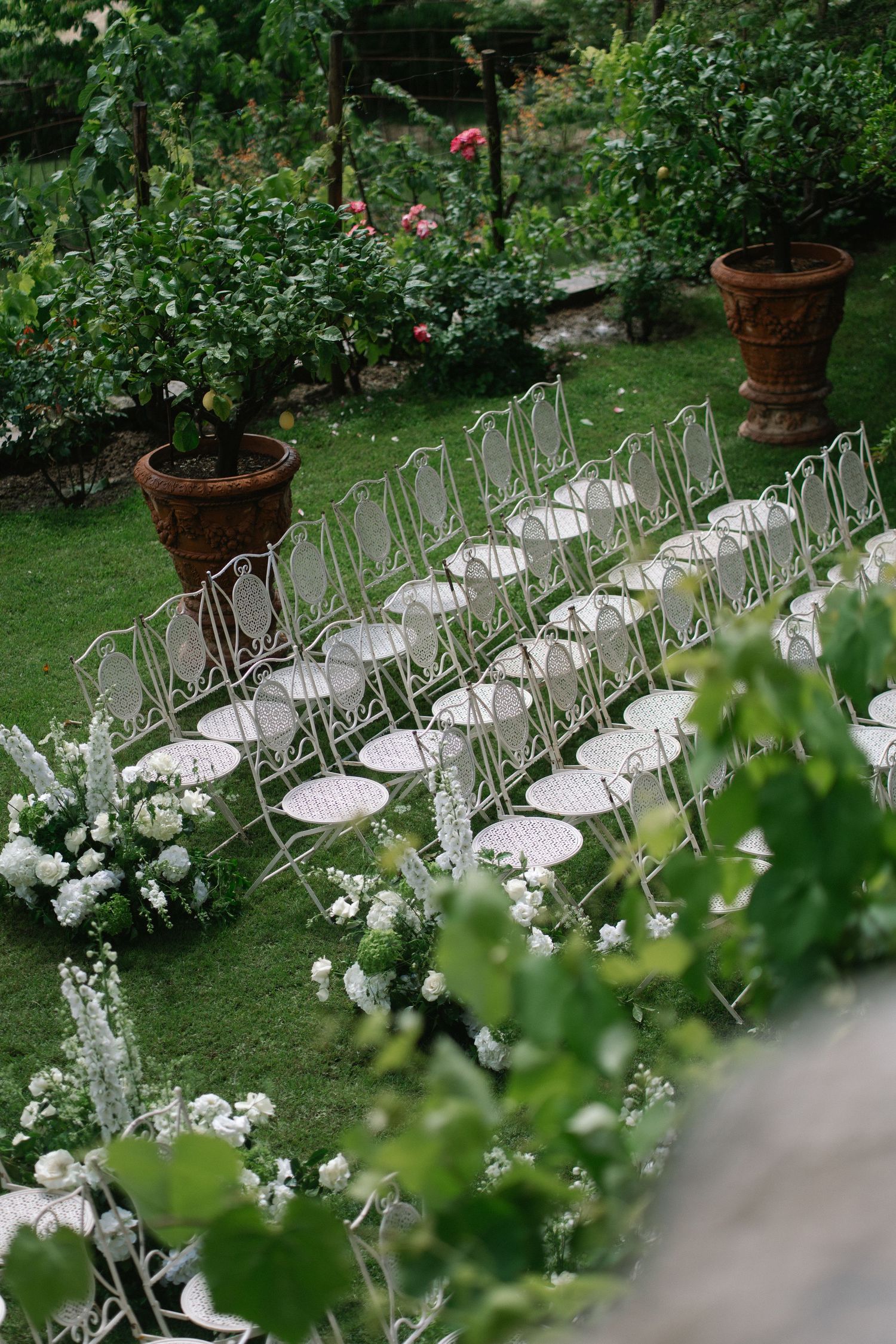 White garden chairs arranged in rows on green grass with floral arrangements and terracotta pots for an outdoor wedding.