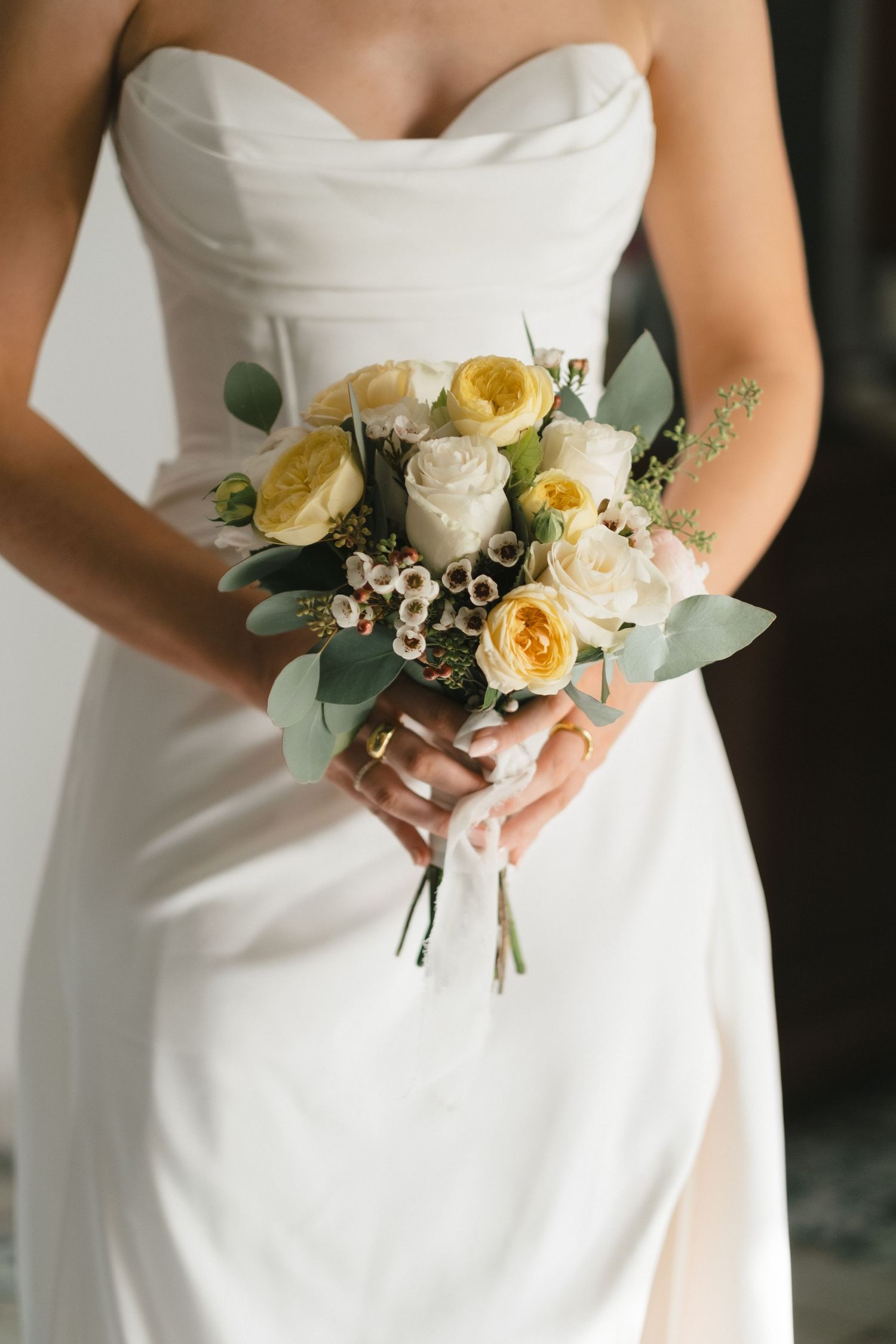 Bride in strapless white gown holding elegant bouquet of cream and yellow roses with eucalyptus greenery.