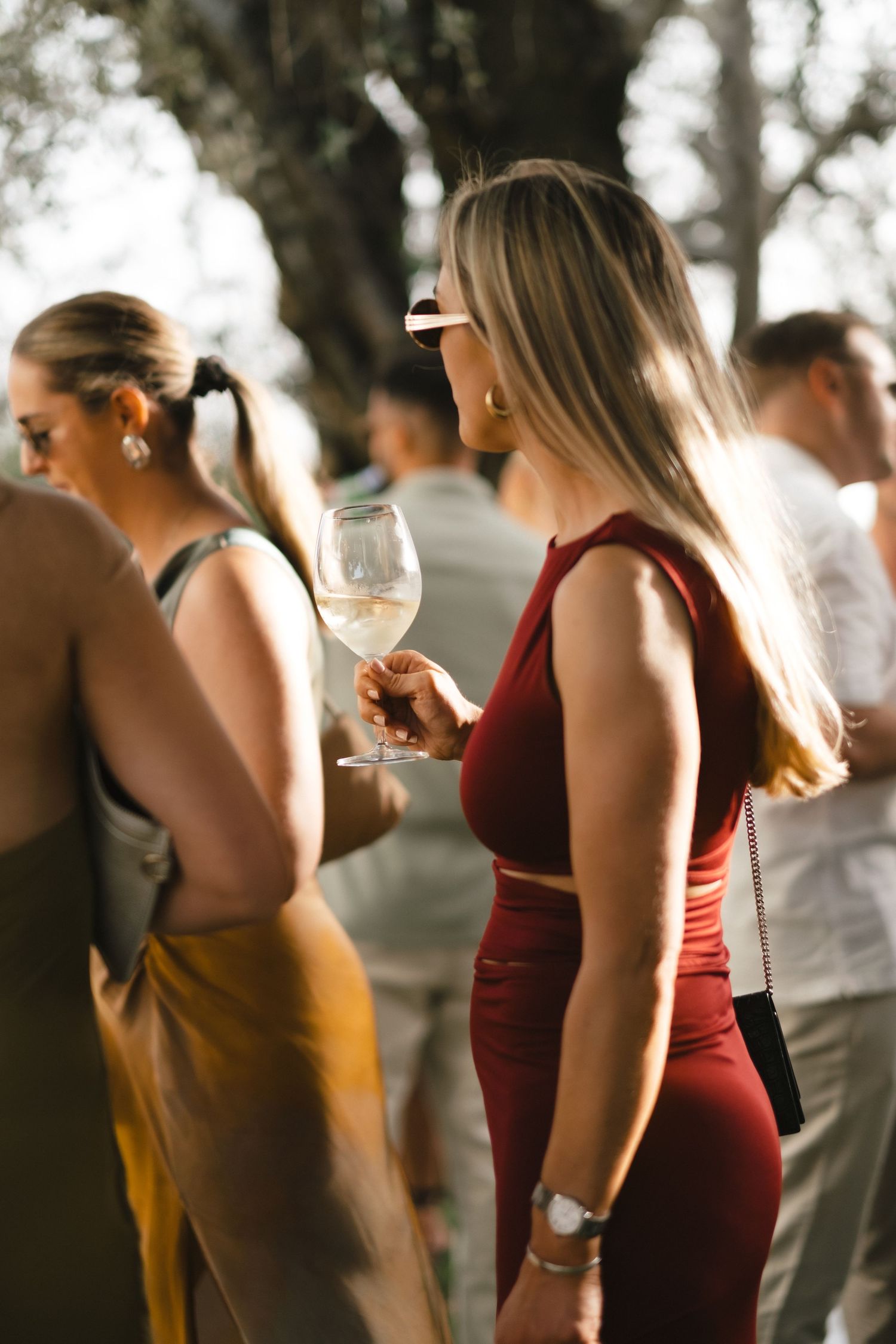 Elegant wedding guest in red dress holds white wine glass at outdoor wedding reception.