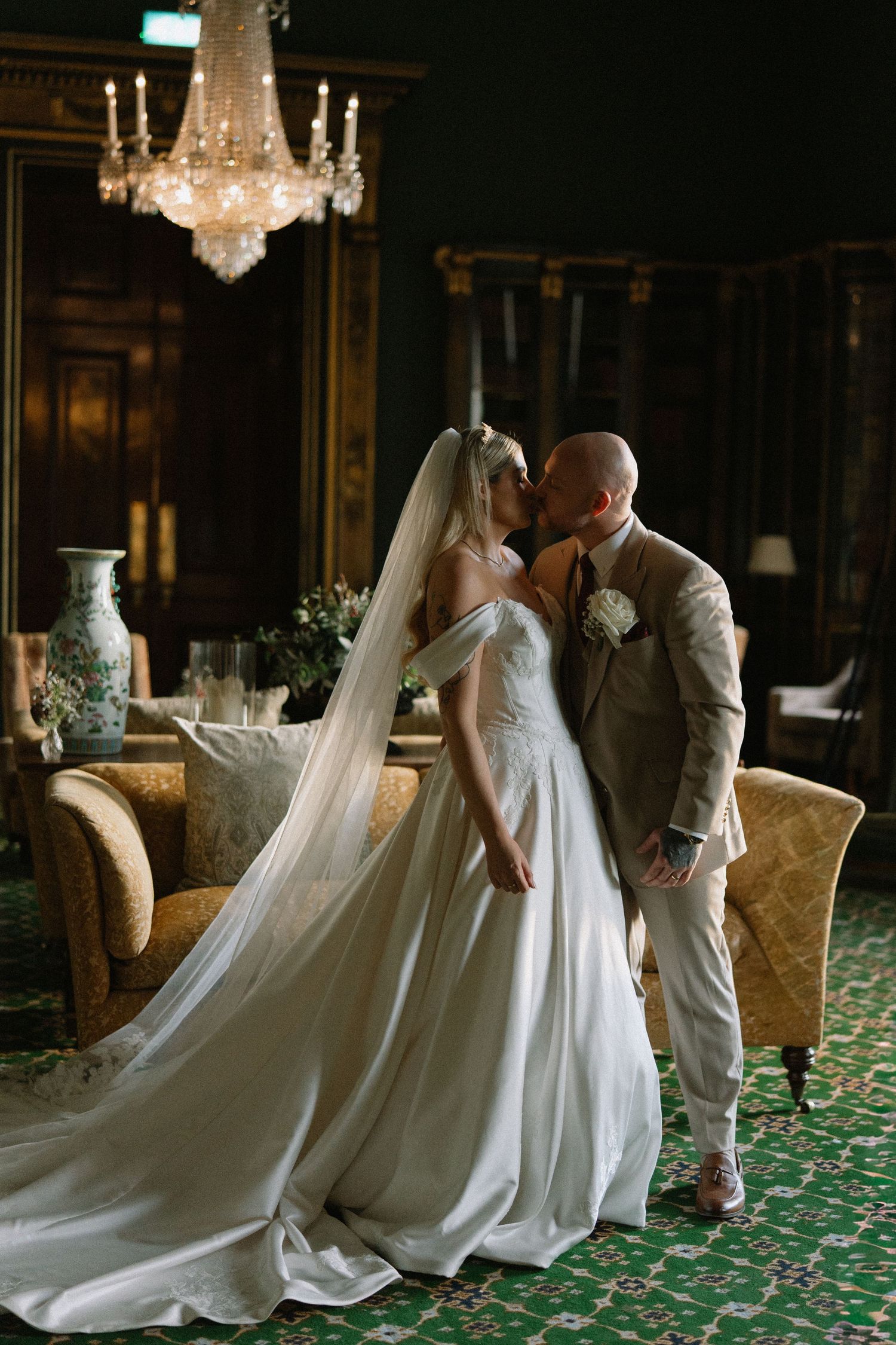 Bride and groom share a romantic kiss in an elegant ballroom with crystal chandelier and green carpet.