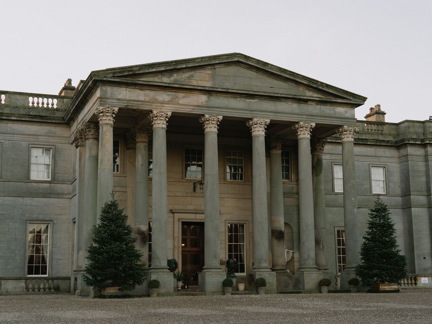 Grand neoclassical mansion with tall columns, symmetrical facade, and evergreen trees flanking the entrance.