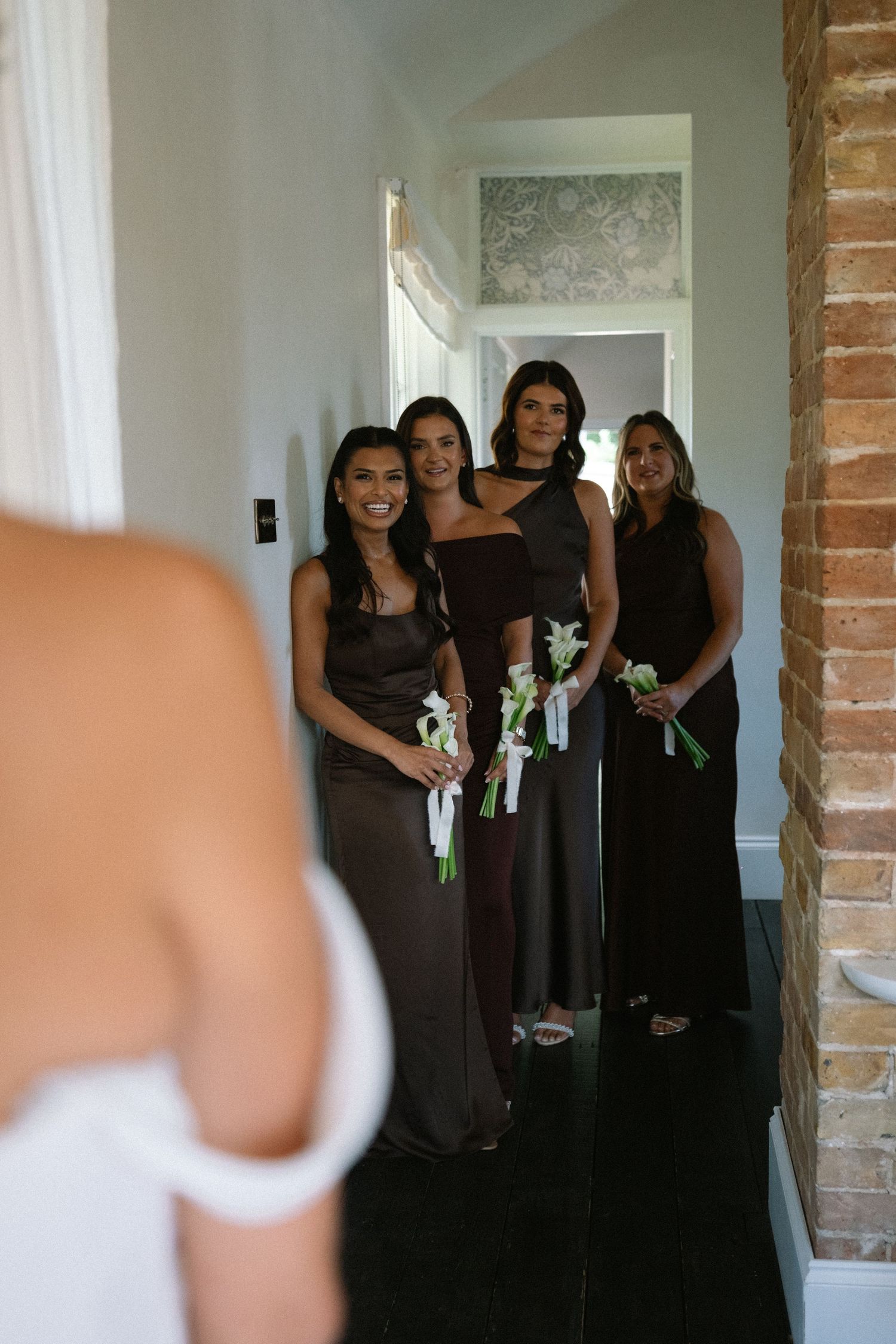 Bridesmaids in dark satin gowns holding white floral boutonnieres react to bride during wedding preparations in hallway.