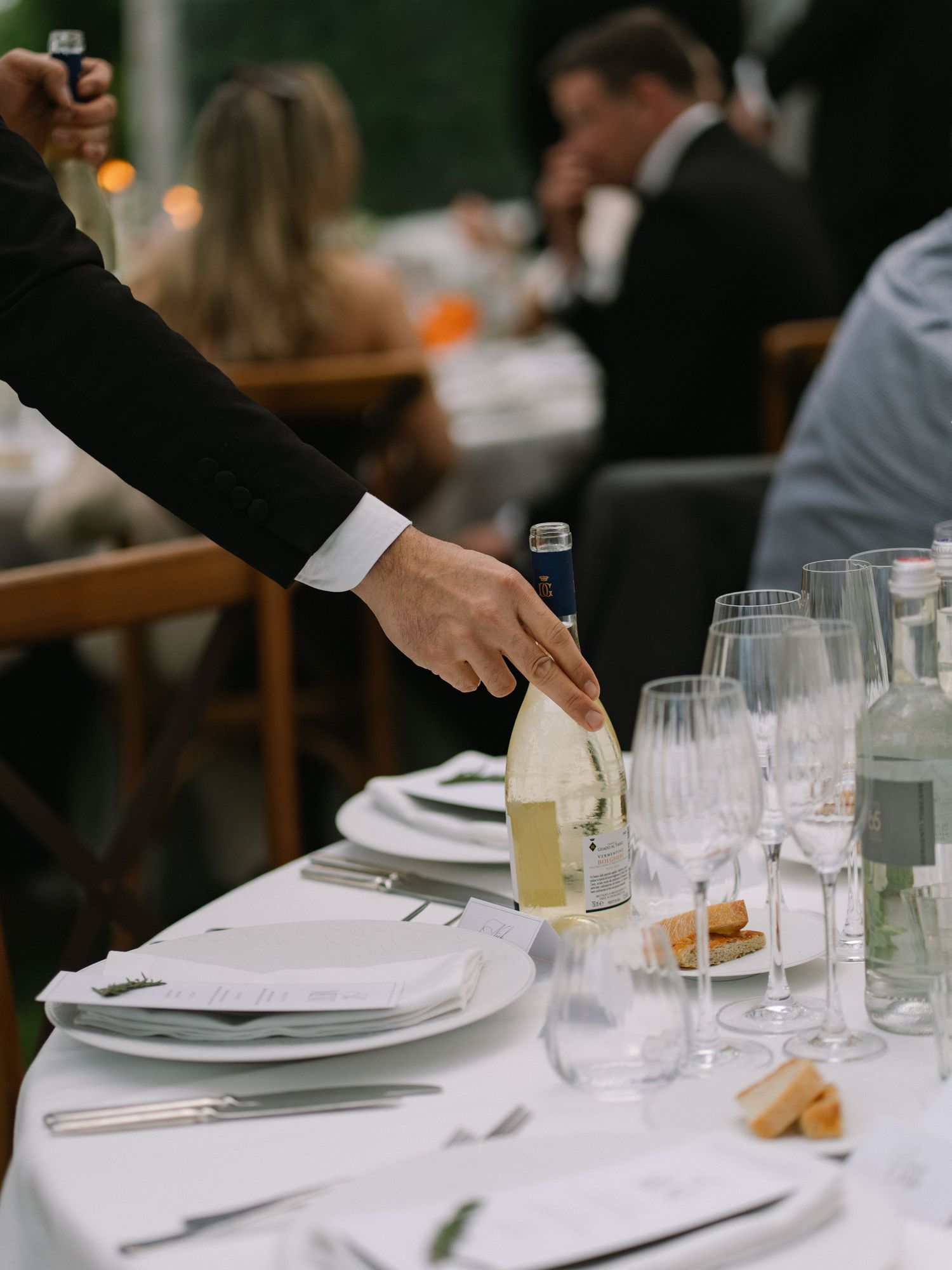 Tuxedo-clad waiter placing a white wine bottle on an elegantly set wedding reception table with crystal glassware.