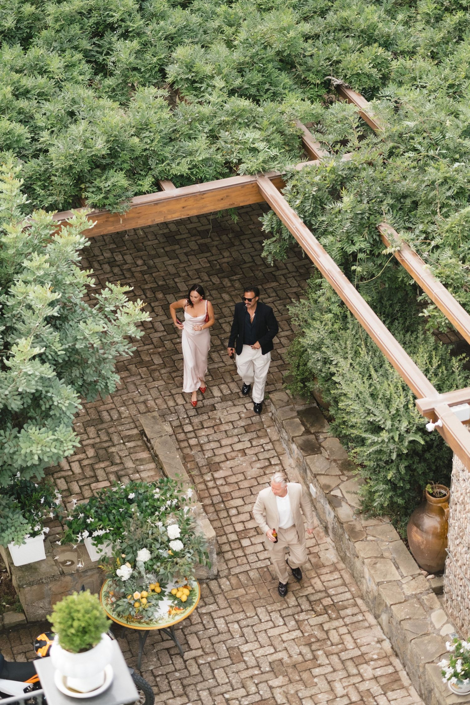 Aerial view of couple walking through lush green garden pergola on brick pathway at outdoor venue.