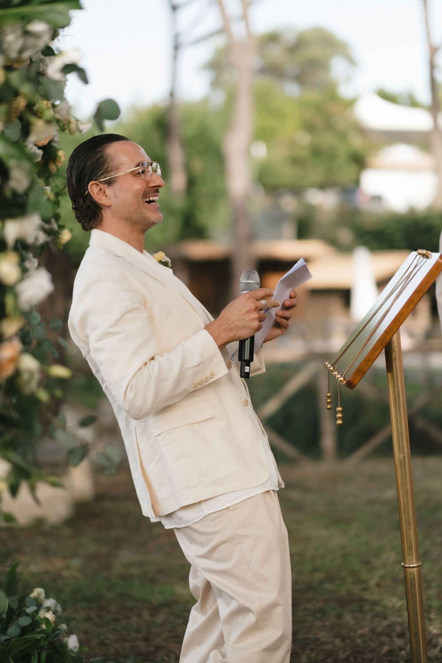 Groom in cream suit laughs while giving speech at outdoor wedding ceremony podium with microphone.