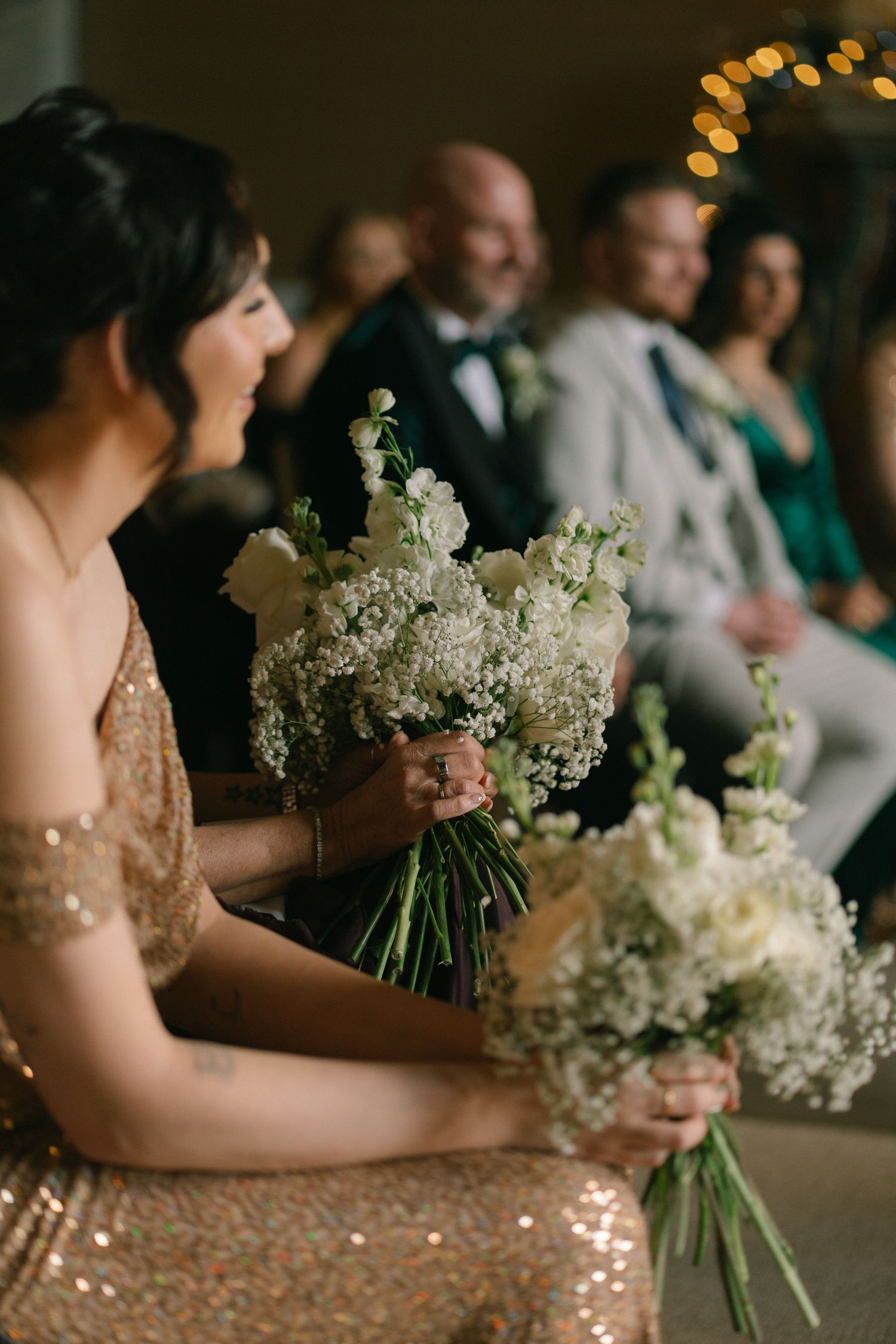 Bridesmaids in gold sequin dresses holding white floral bouquets during a wedding ceremony with guests seated in background.