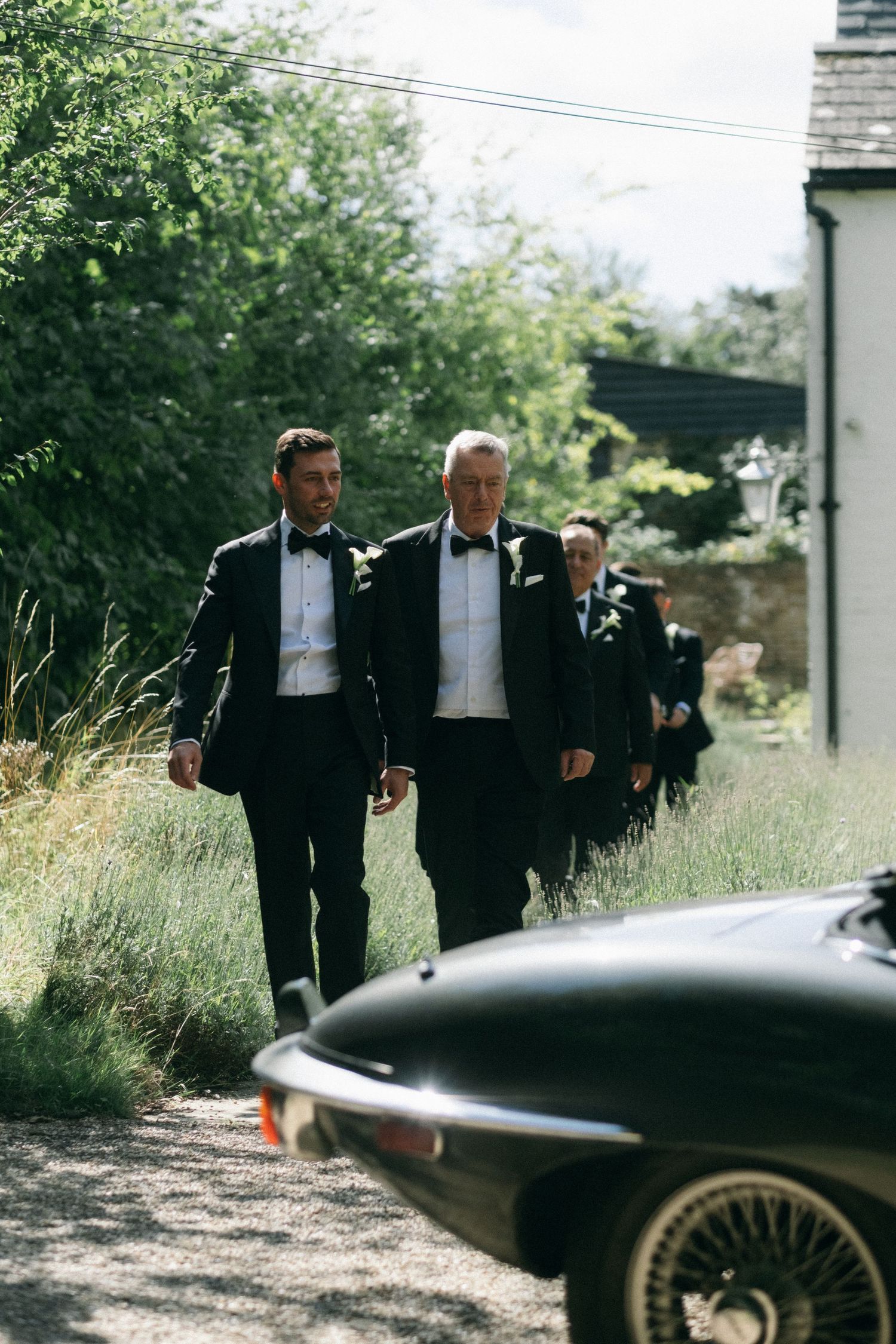 Groomsmen in black tuxedos walking toward a vintage car at an outdoor wedding venue.
