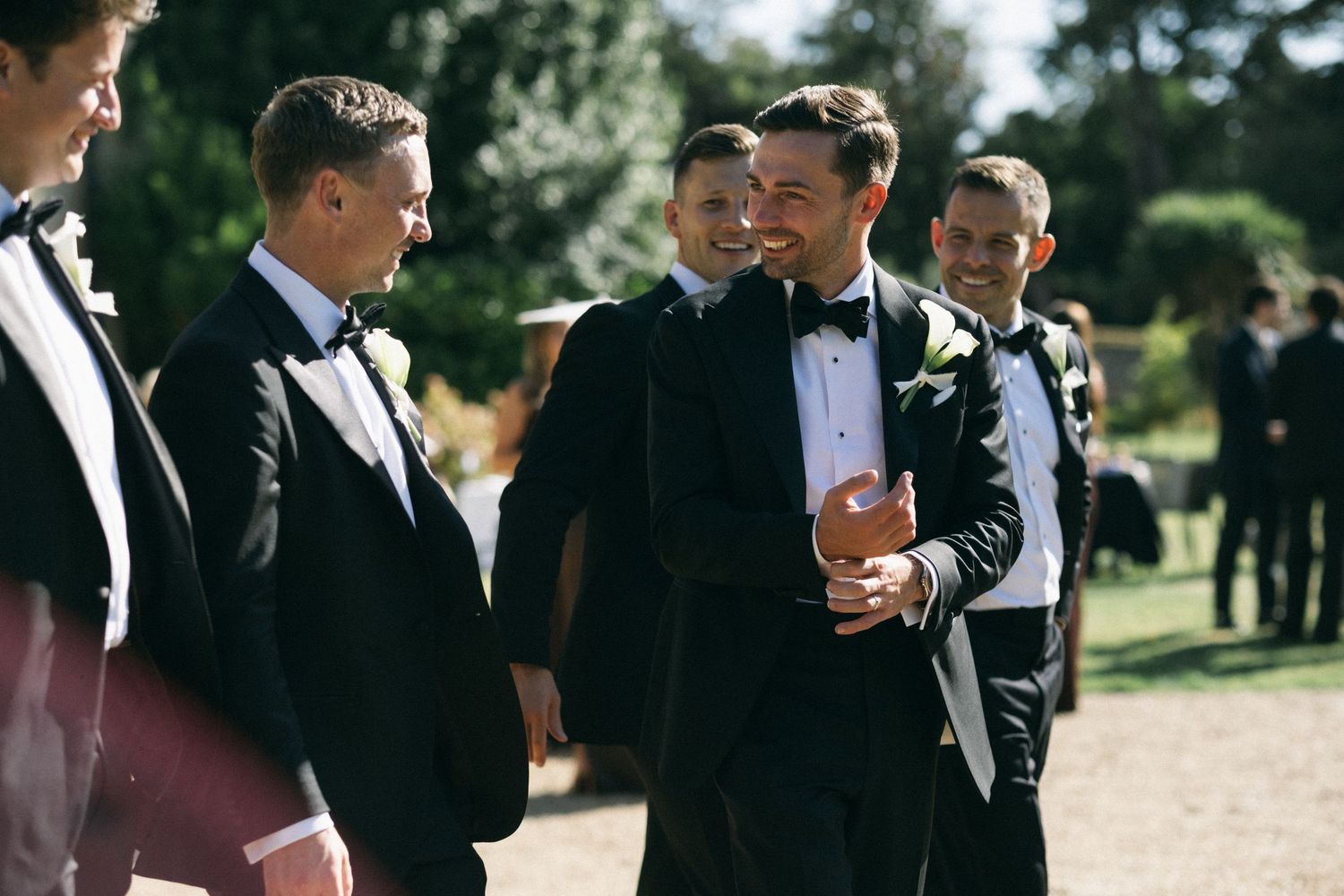 Groomsmen in black tuxedos with bow ties laughing and socializing outdoors at a wedding reception.