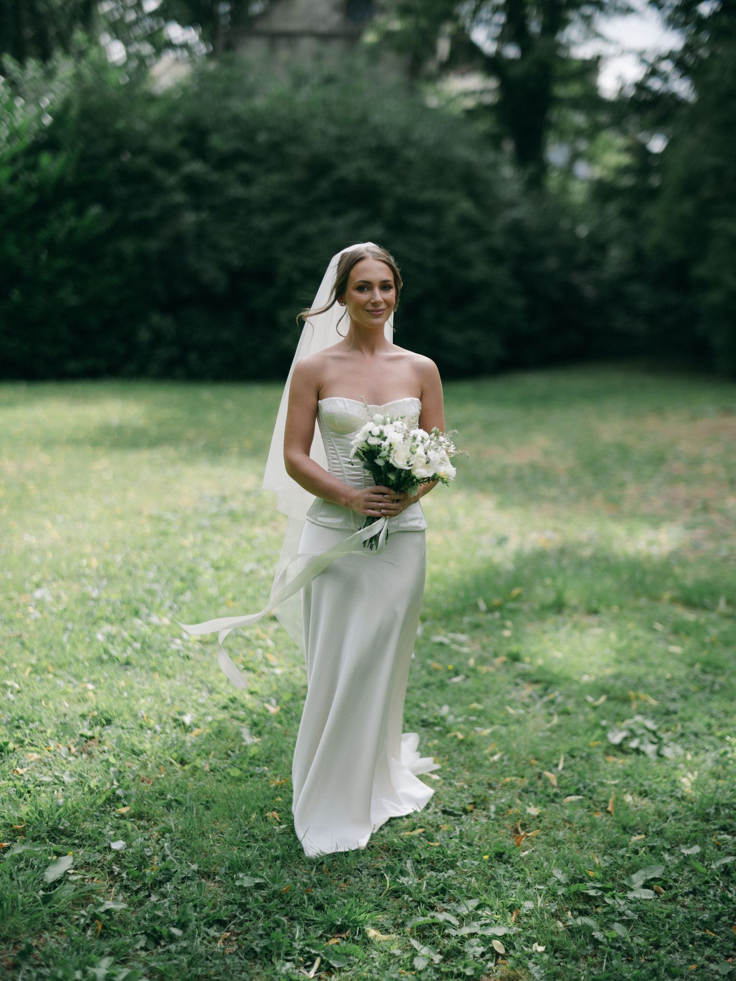Smiling bride in strapless white gown holding bouquet of white flowers, walking on green grass with trees in background.