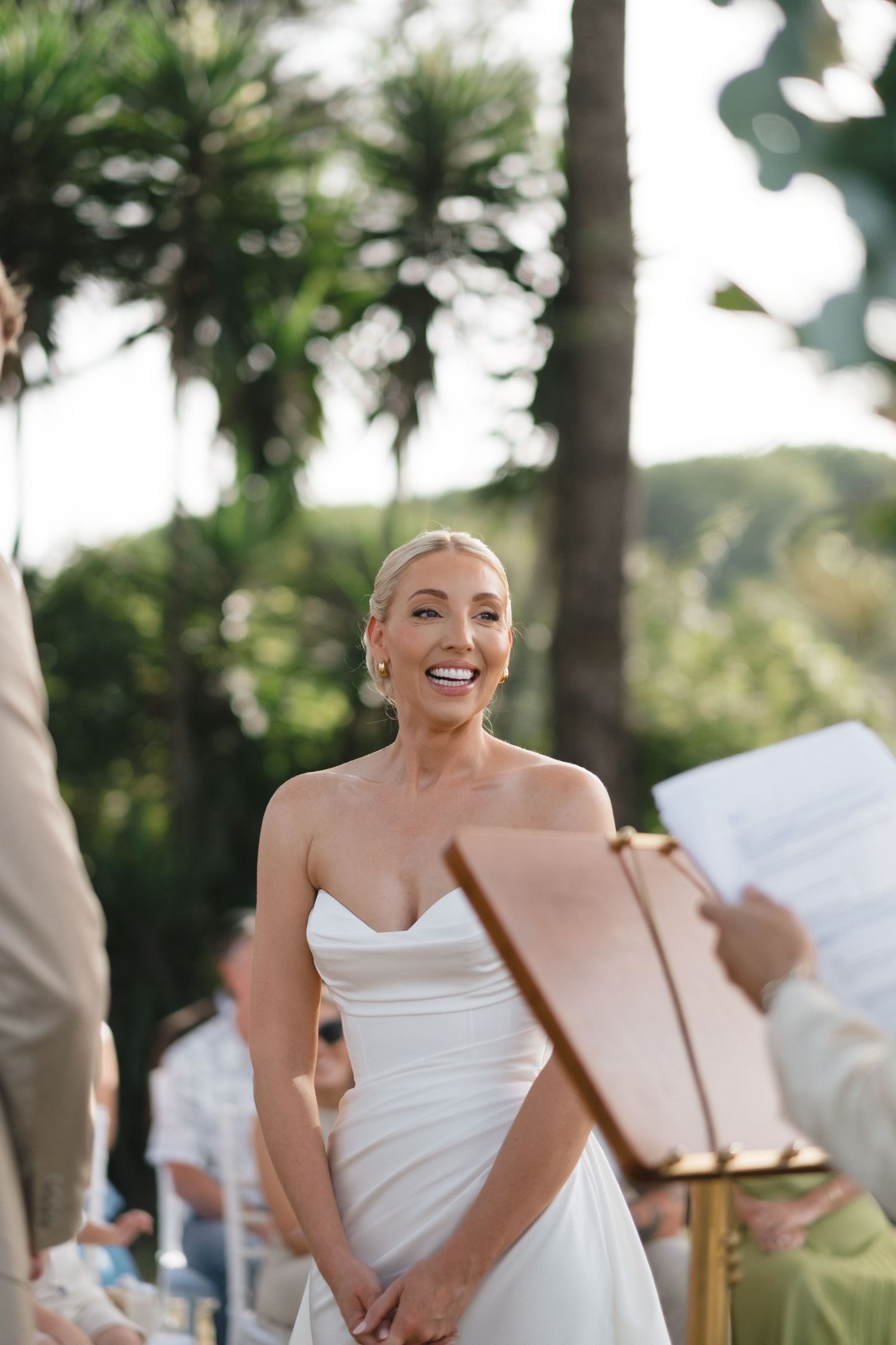 Smiling bride in strapless white gown during outdoor wedding ceremony with lush green palm trees in background.