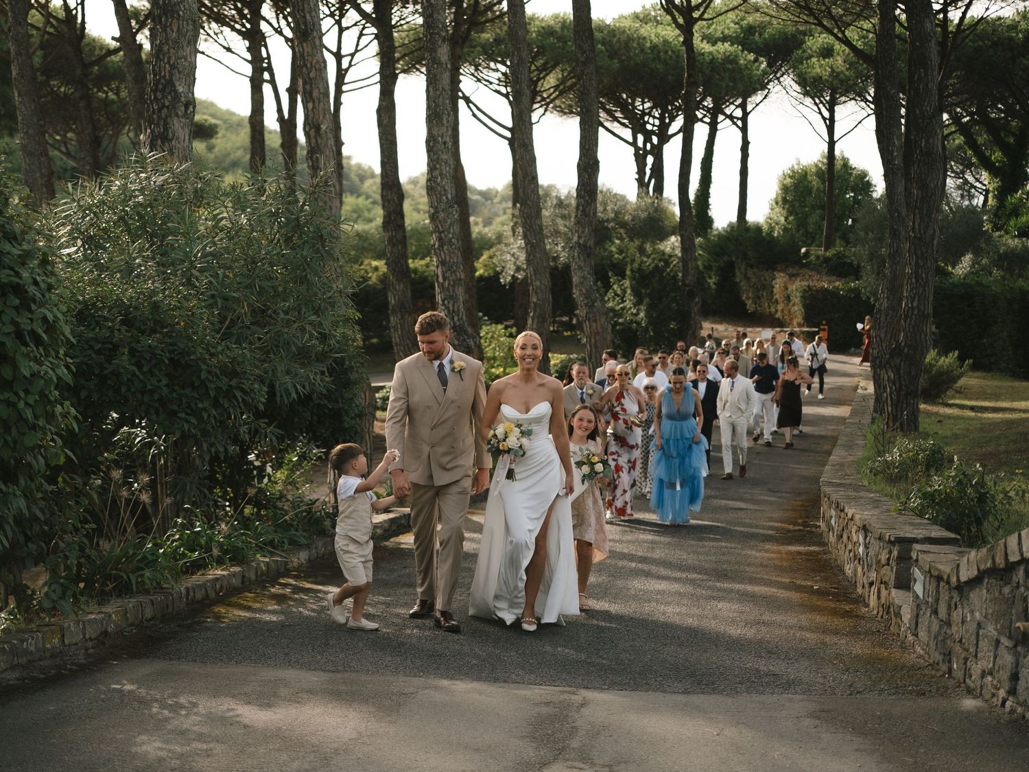 Wedding procession walking down a tree-lined path, bride in white gown with groom and guests following.