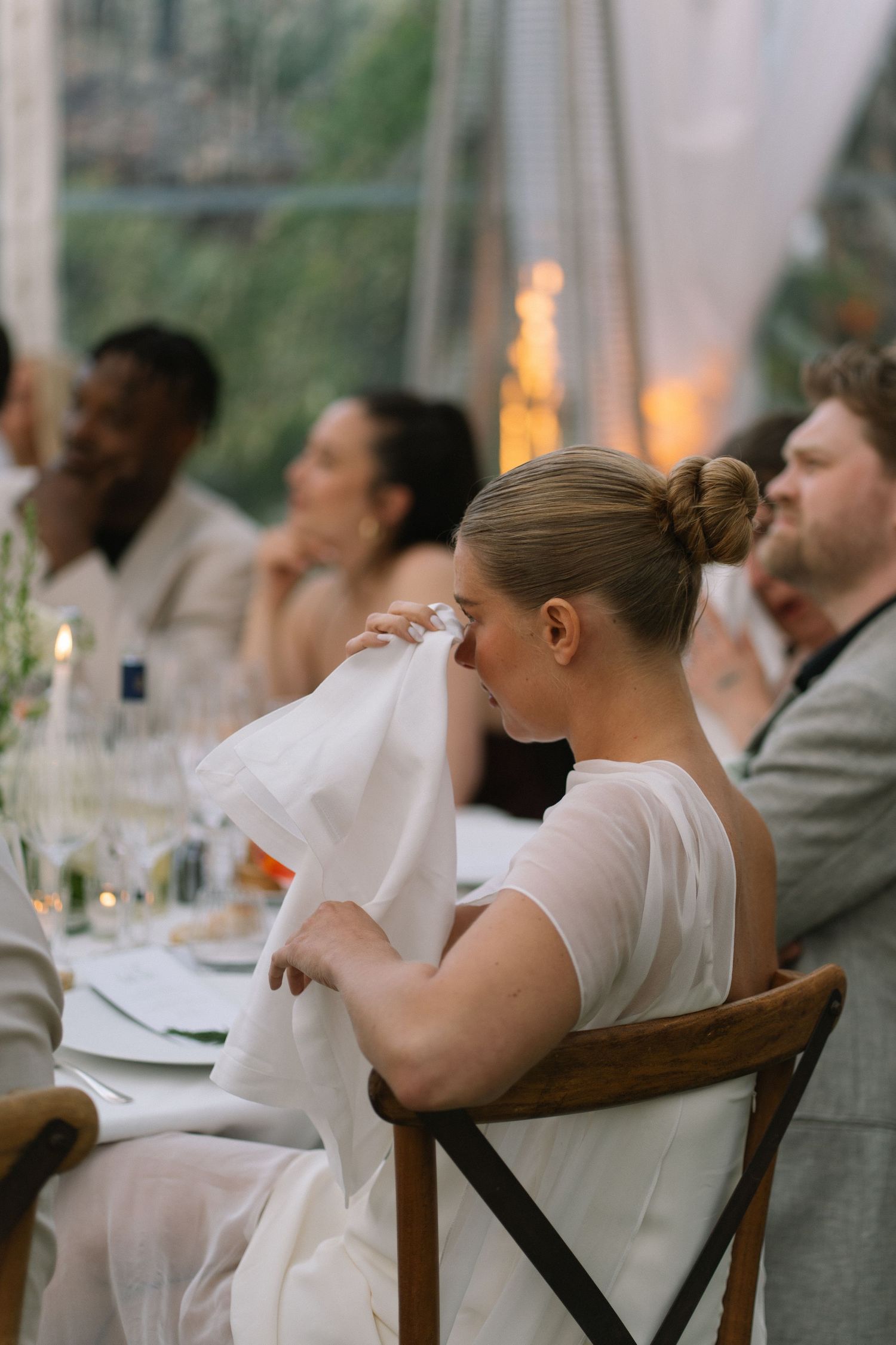 Emotional bride wiping tears with white napkin during heartfelt wedding reception speech at candlelit dinner table.