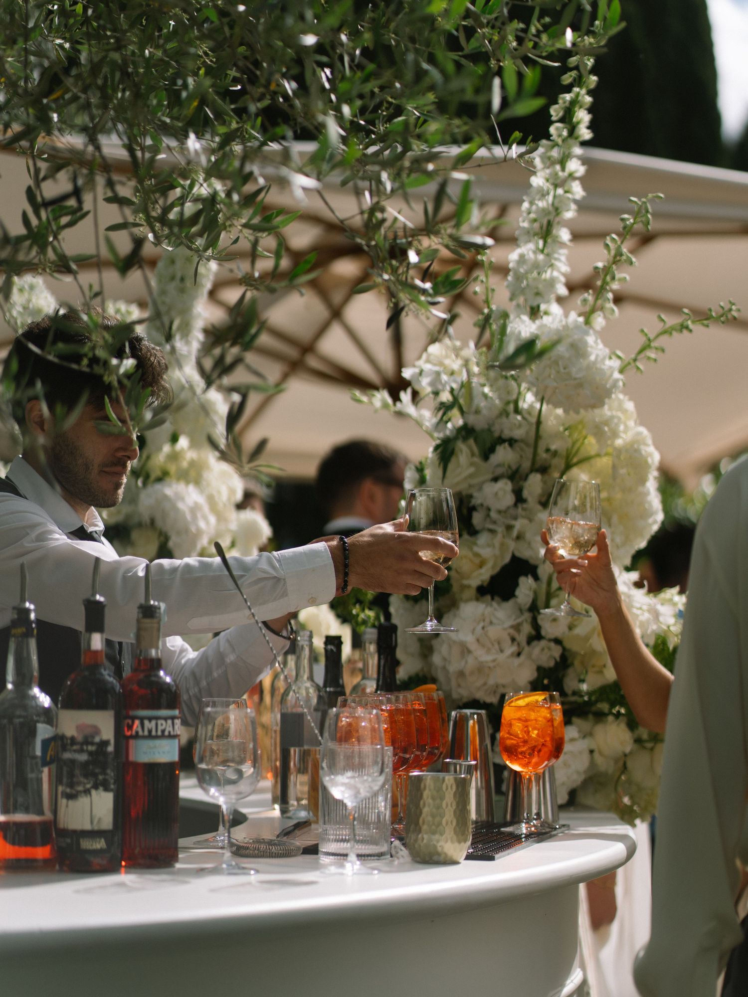 Bartender serves Campari cocktails at an elegant outdoor wedding bar adorned with white floral arrangements.