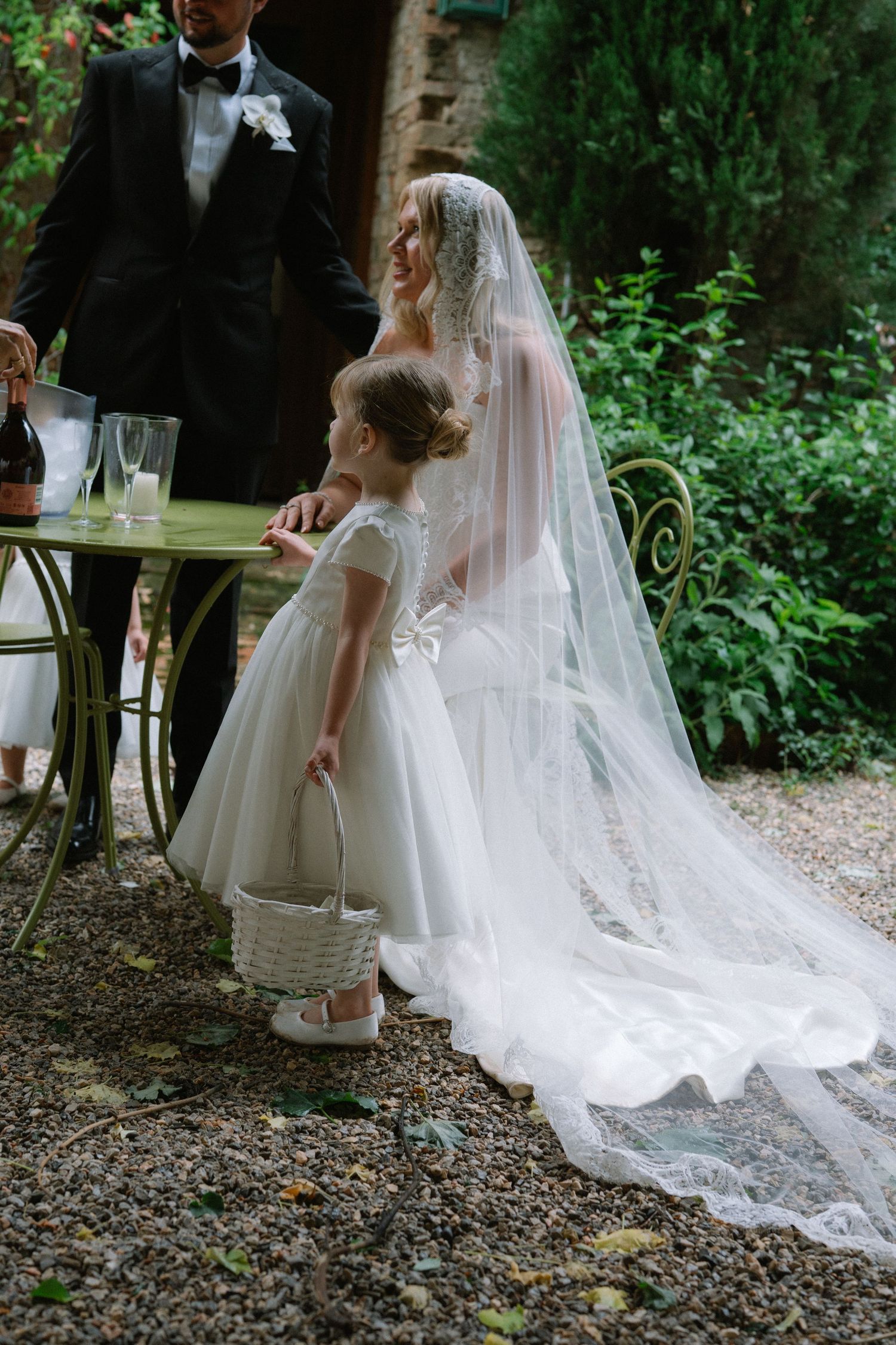 Young flower girl in white dress stands beside bride in lace veil at outdoor garden reception table.