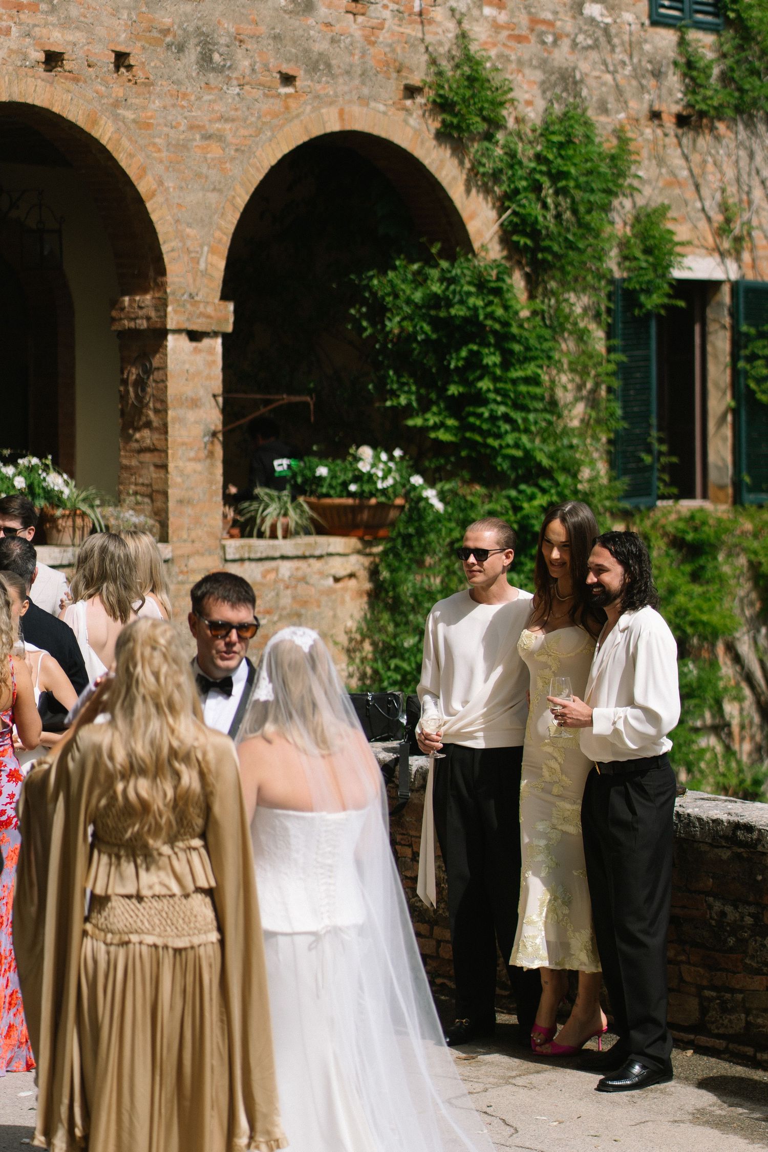 Bride in a white gown and veil greeting wedding guests under stone arches of a Tuscan villa courtyard.