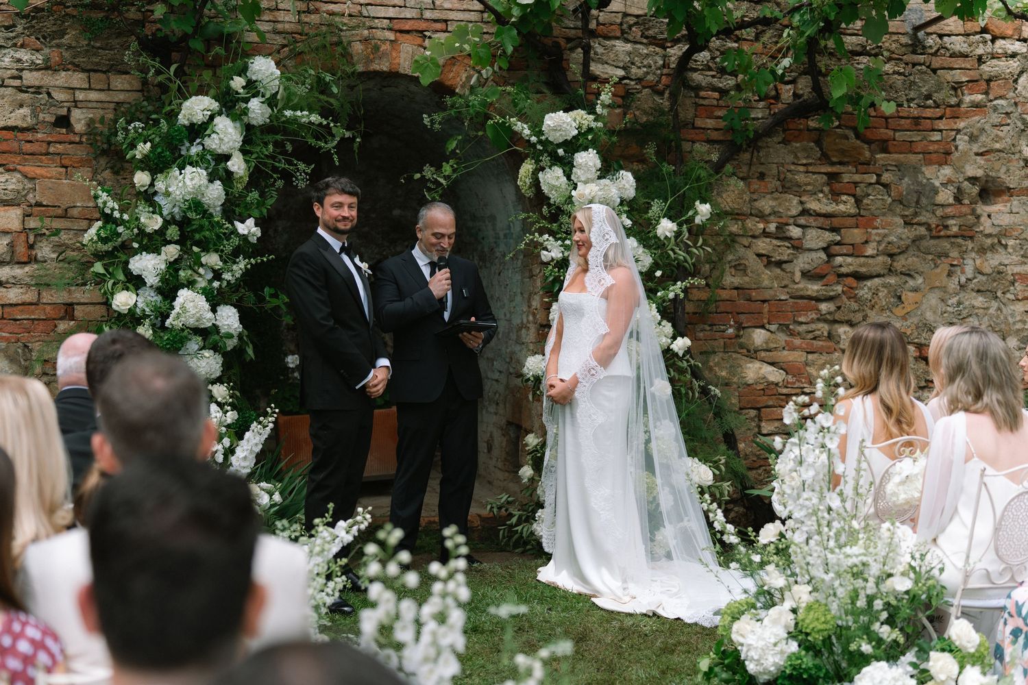 Bride and groom stand at the altar beneath a lush white floral arch during outdoor wedding.