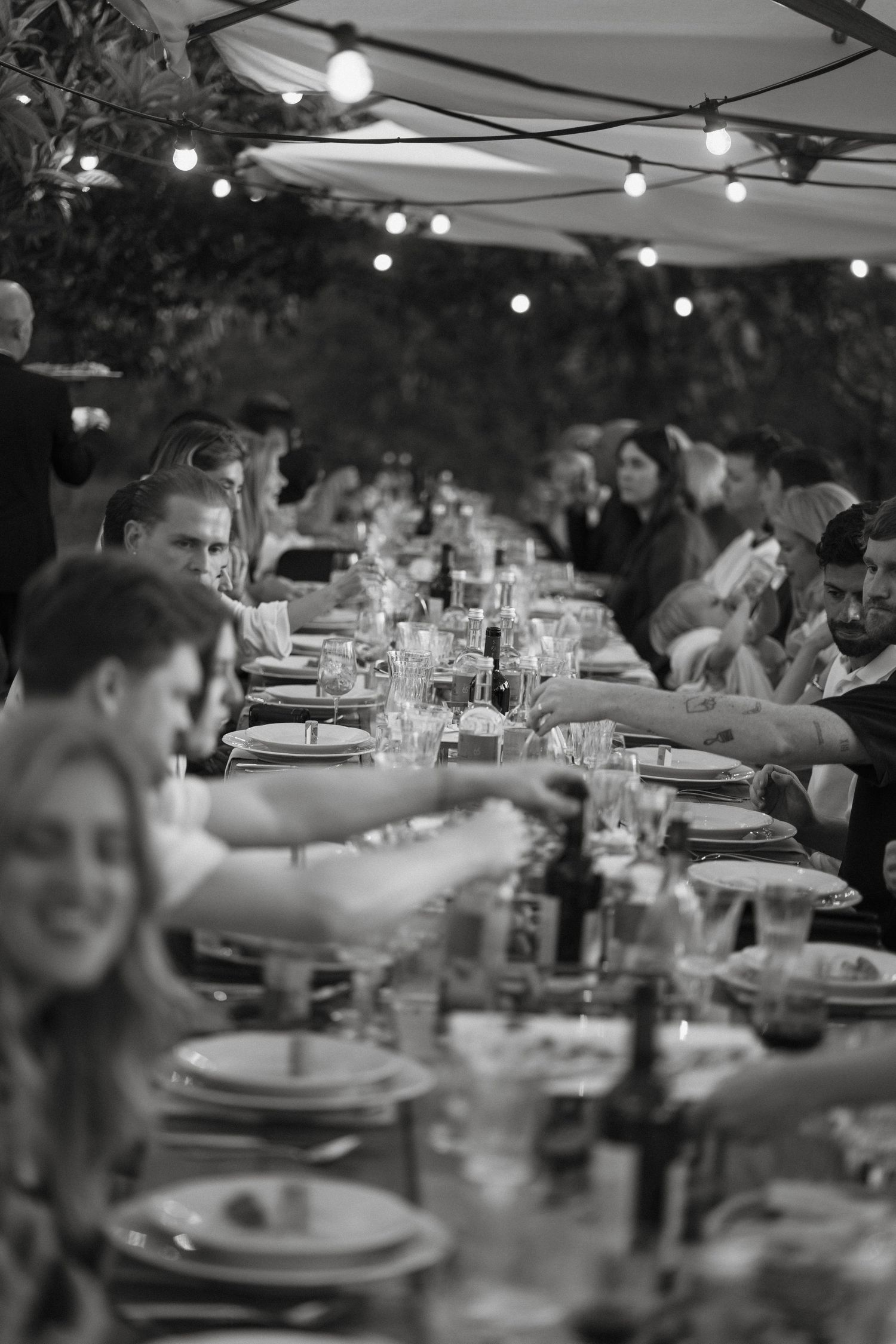 Black and white photo of guests laughing and dining at a long festive table adorned with string lights.
