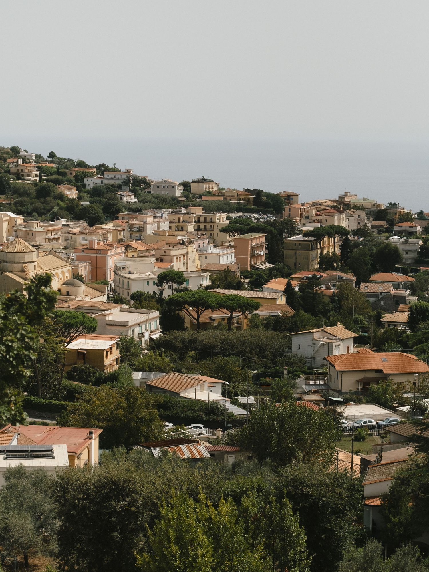Aerial view of a picturesque Italian hillside town with terracotta rooftops, lush trees, and the sea in the background.