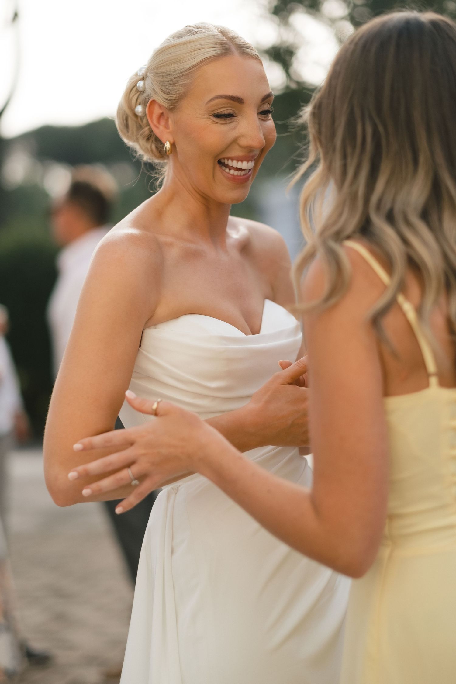 Smiling bride in strapless white gown laughing with guest at outdoor wedding reception.