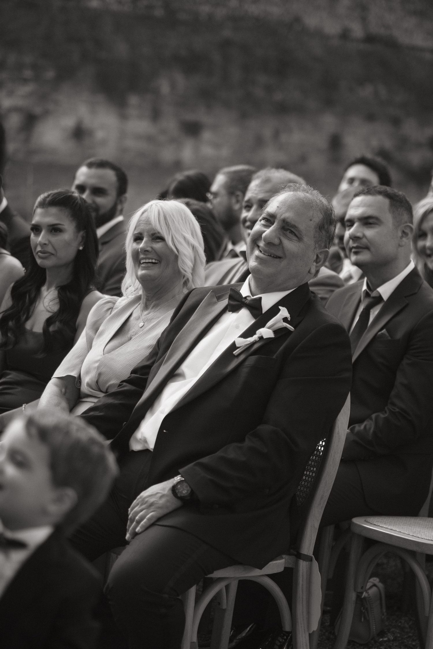 Black and white photo of smiling wedding guests seated at an outdoor ceremony, man in tuxedo with bow tie in foreground.