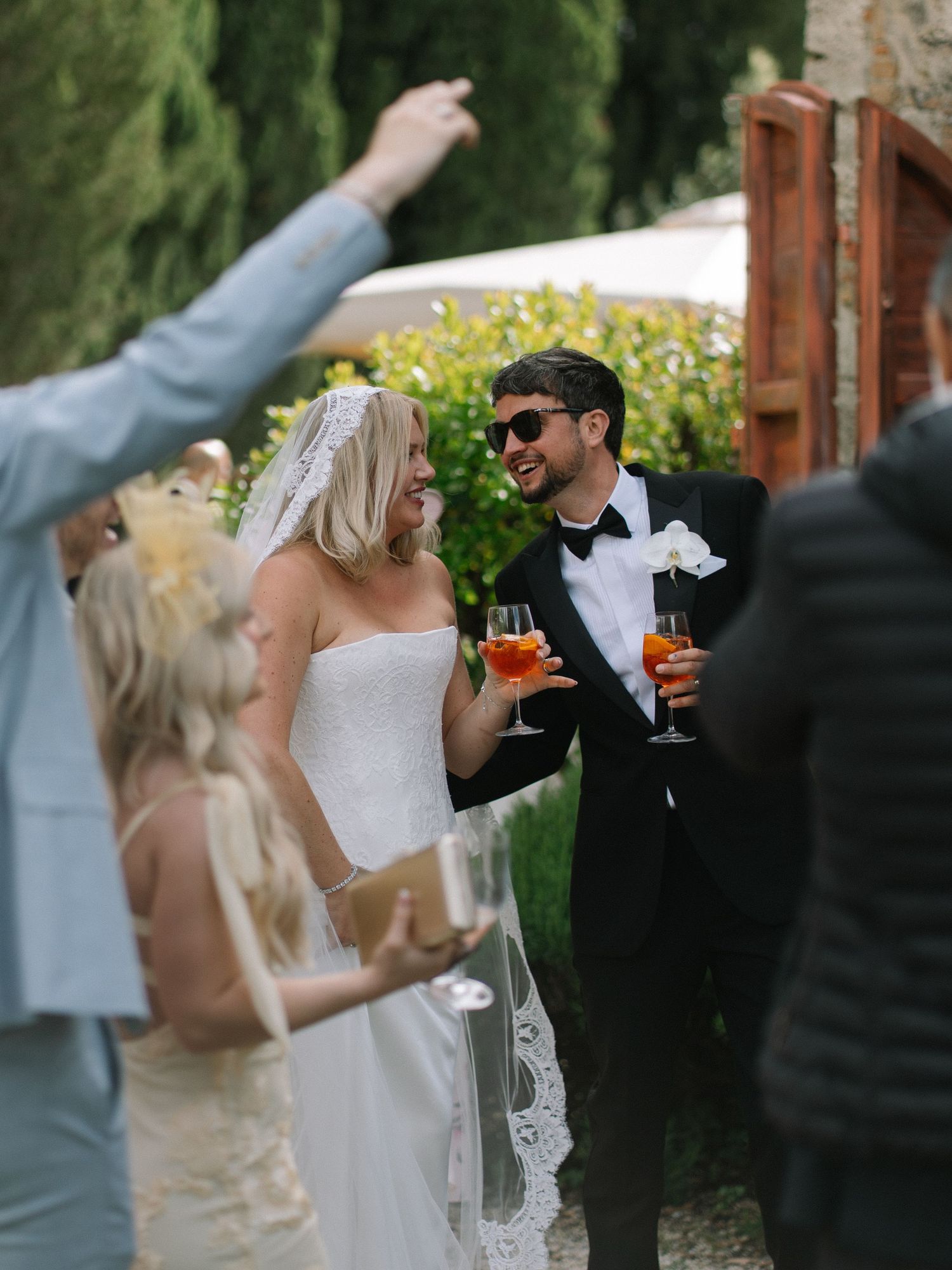 Bride and groom smiling and holding Aperol Spritz drinks at an outdoor wedding reception with guests.